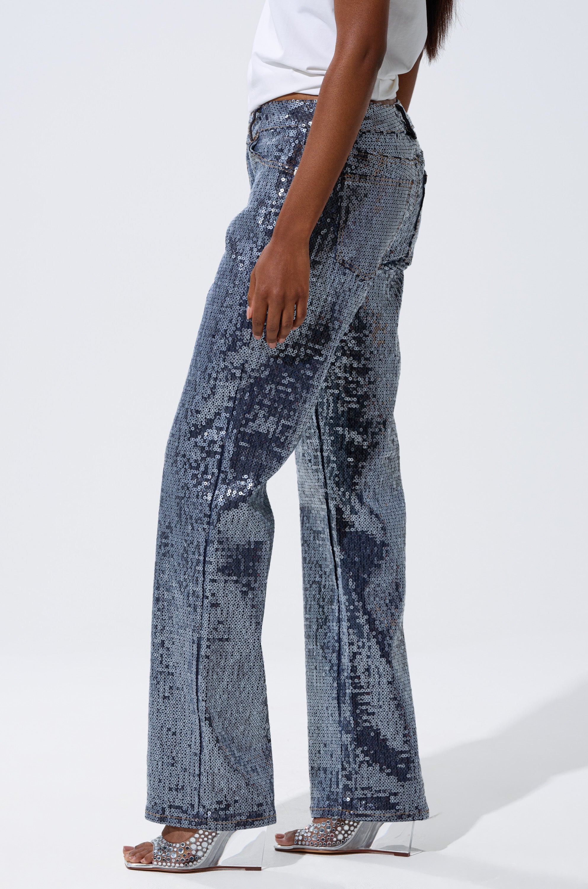 A person stands in profile against a plain light background wearing the ASHTON SEQUIN DENIM PANT, a white top, and open-toe heeled sandals.