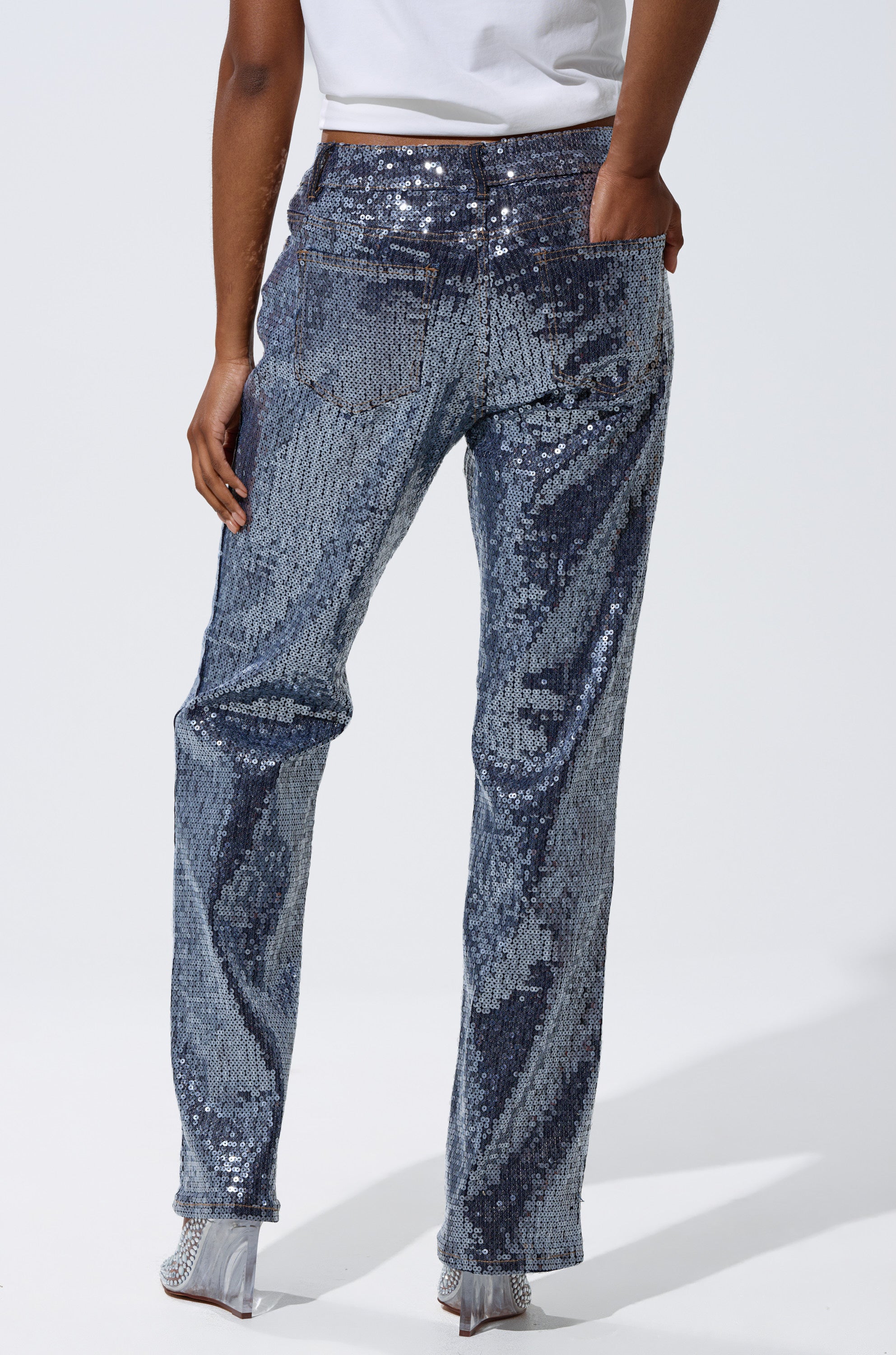 A person stands facing away, wearing a white shirt, silver high-heeled shoes, and the ASHTON SEQUIN DENIM PANT against a plain light background.