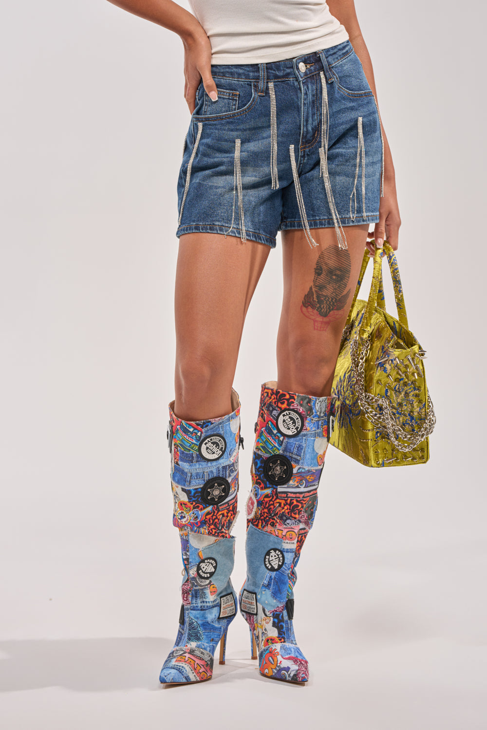 Wearing the CAME THROUGH DRIPPIN DENIM SHORT with white trim, a white tank top, colorful patchwork knee-high boots, and holding a shiny yellow patterned handbag. A tattoo is visible on their thigh—the ultimate festival look. 1
