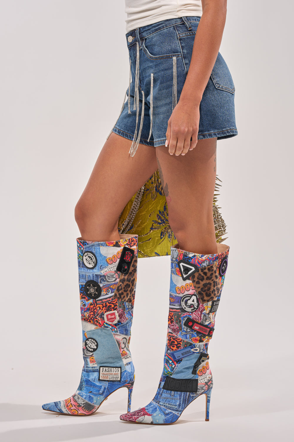 A person wearing the CAME THROUGH DRIPPIN DENIM SHORT and colorful knee-high boots with patchwork prints (leopard, text, graphics) poses against a plain background, showcasing a bold festival outfit. 3