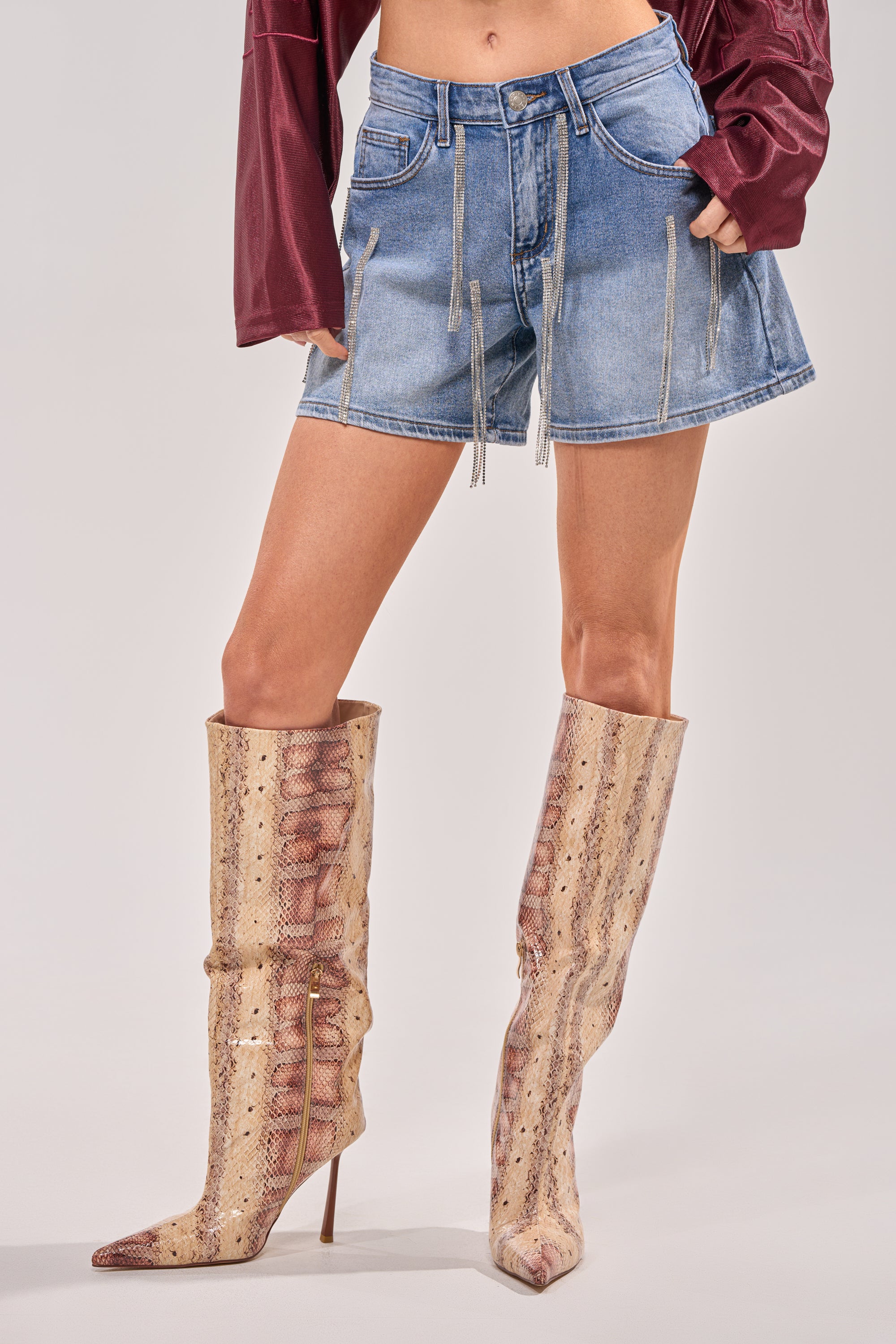 A person showcases festival style in a burgundy long-sleeve top, DRIP CITY DENIM SHORT blue high-waist zipper-detailed shorts, and knee-high pointed-toe snakeskin boots against a light background. 0