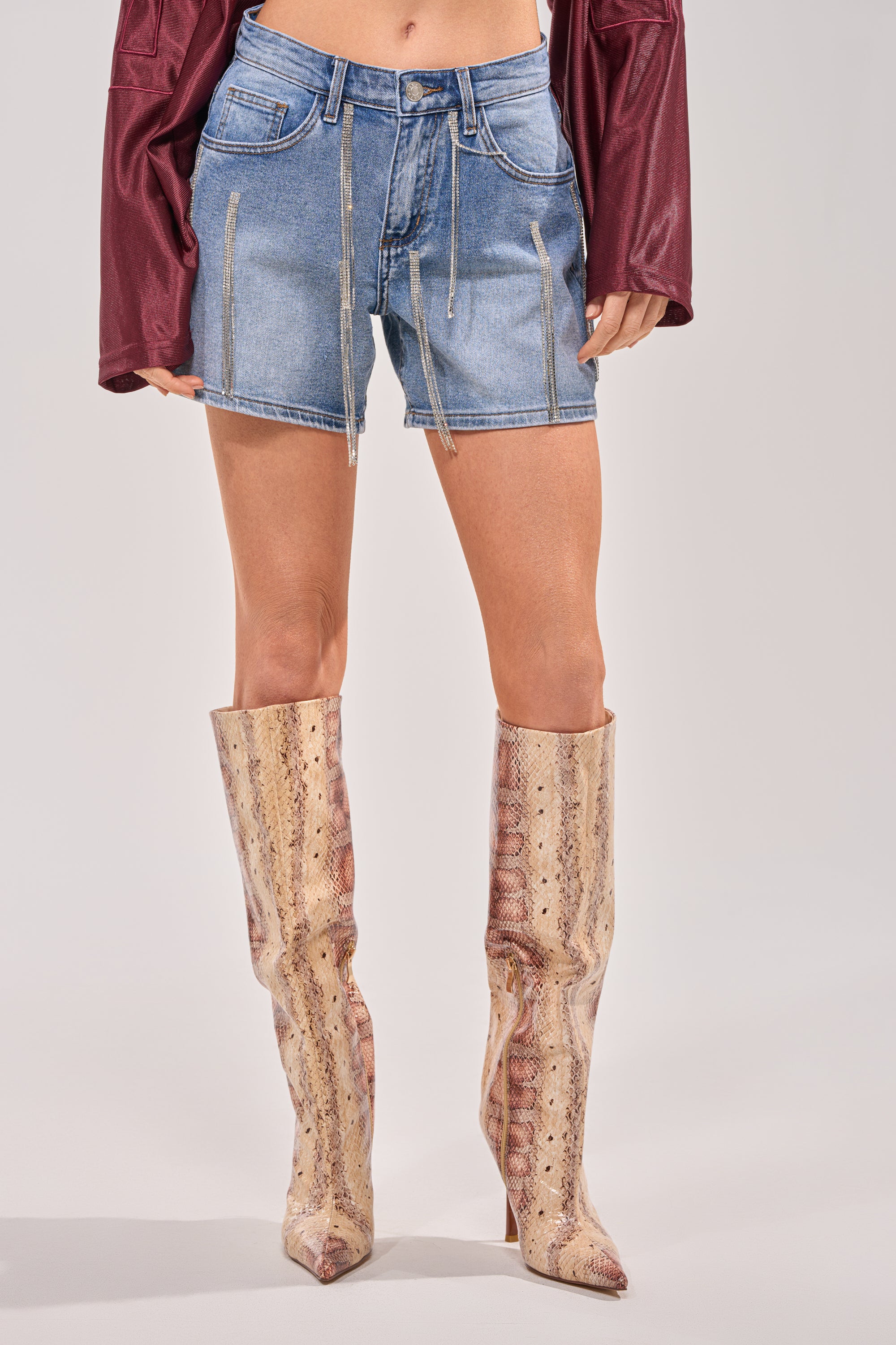 Wearing DRIP CITY DENIM SHORTS, a maroon long-sleeve top, and tall pointed-toe tan snakeskin boots, this look stands out against a plain backdrop—ideal for festival-ready style. 1
