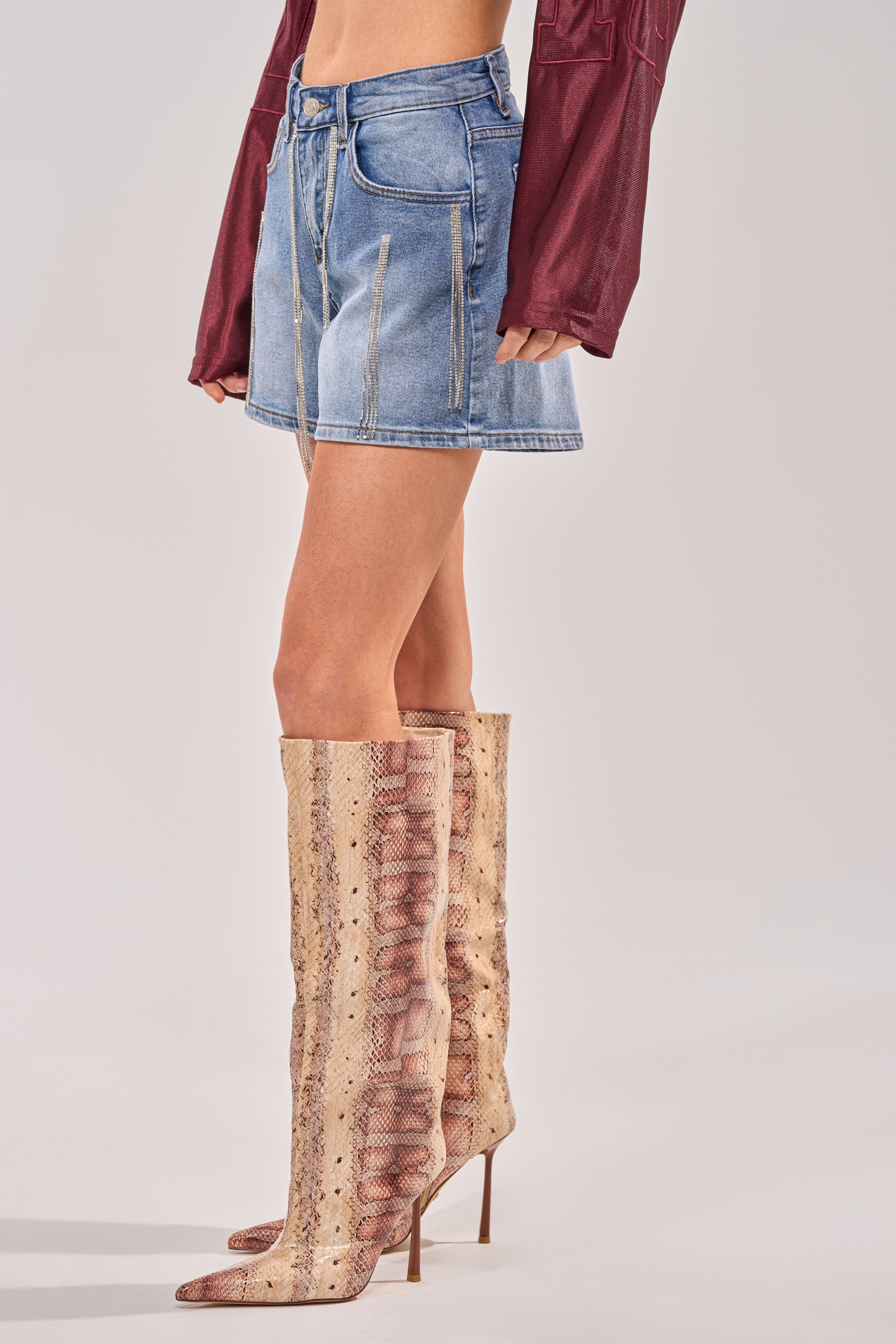 A person wears the DRIP CITY DENIM SHORT, a maroon long-sleeve top, and knee-high pointed-toe snakeskin boots—perfect festival style inspiration set against a plain background. 2