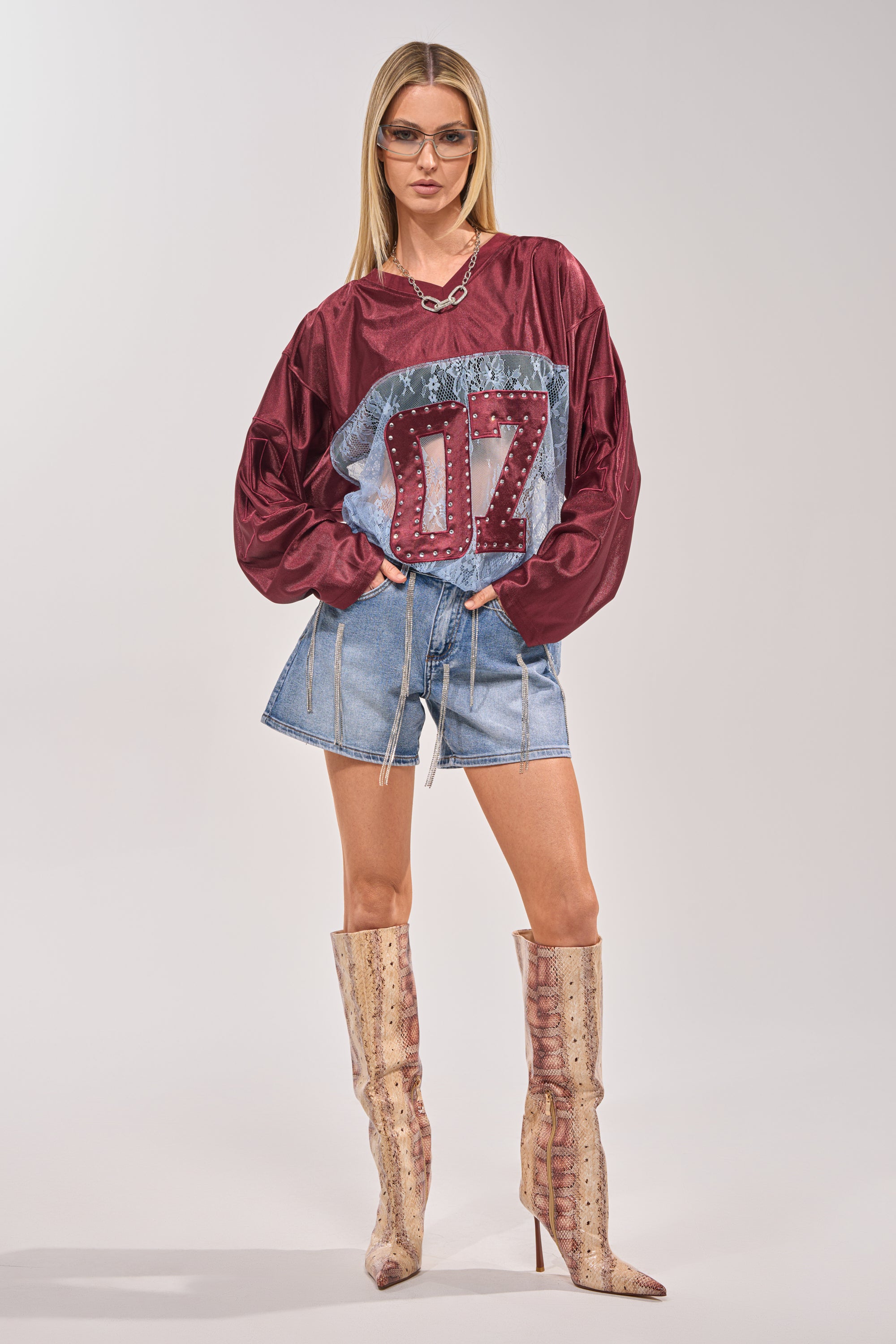 A woman wearing glasses, a maroon "07" sweater, DRIP CITY DENIM SHORT high-rise light blue shorts with zippers, and tall beige patterned boots poses with hands on hips against a plain background, showcasing festival style. 5