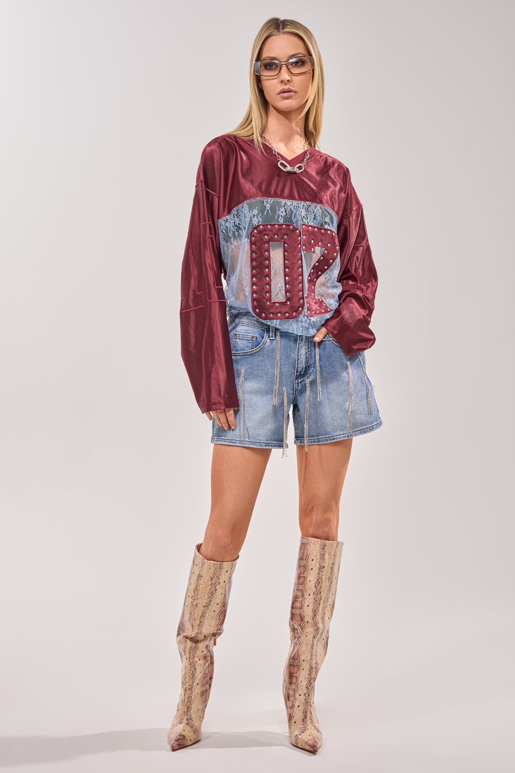 A woman exudes confidence in a maroon sports jersey, DRIP CITY DENIM SHORT, patterned tall boots, sunglasses, and a chunky necklace against a simple light background. 6