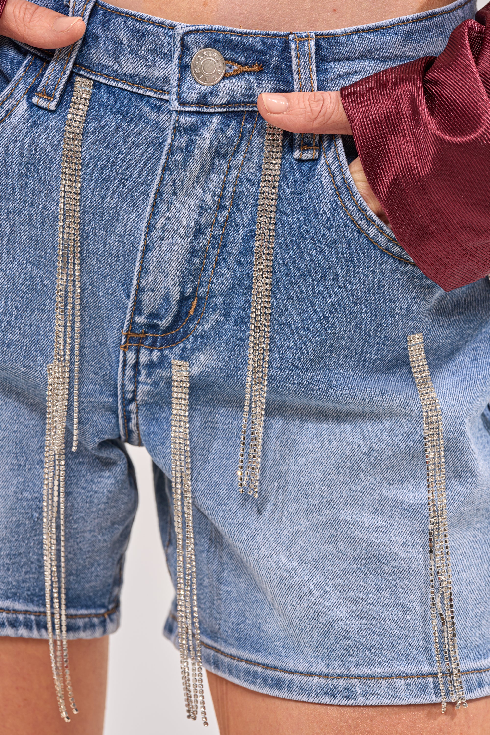 Close-up of DRIP CITY DENIM SHORT featuring rhinestone fringe, a high-rise waist, and silver chain details. Styled with a burgundy long-sleeve top for standout festival looks; hand rests in the pocket for added flair. 7