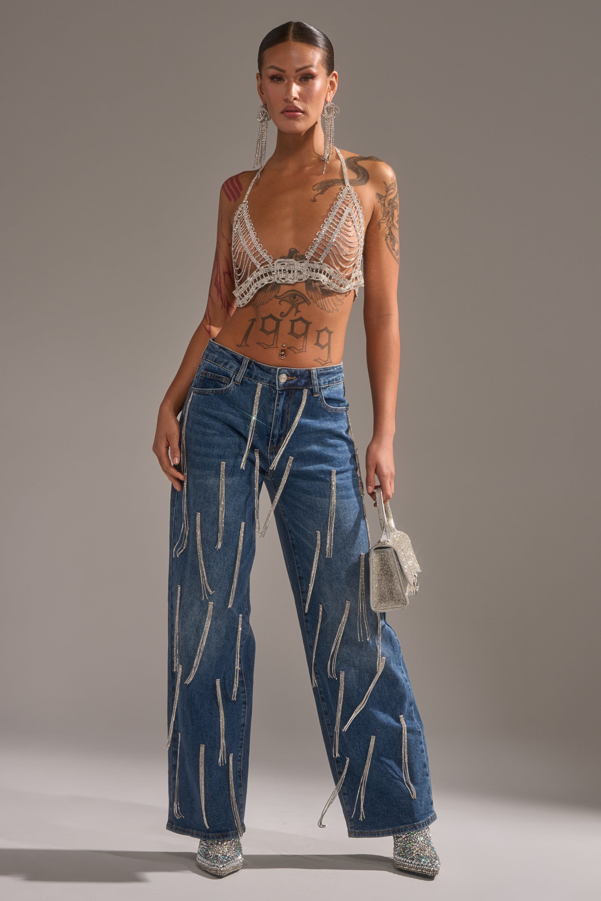 A woman faces forward wearing the DRIPPED OUT RELAXED DENIM wide-leg jeans with zipper details, styled with a beaded halter top, sparkly heels, long earrings, a glittery bag, visible tattoos, and her hair pulled back.