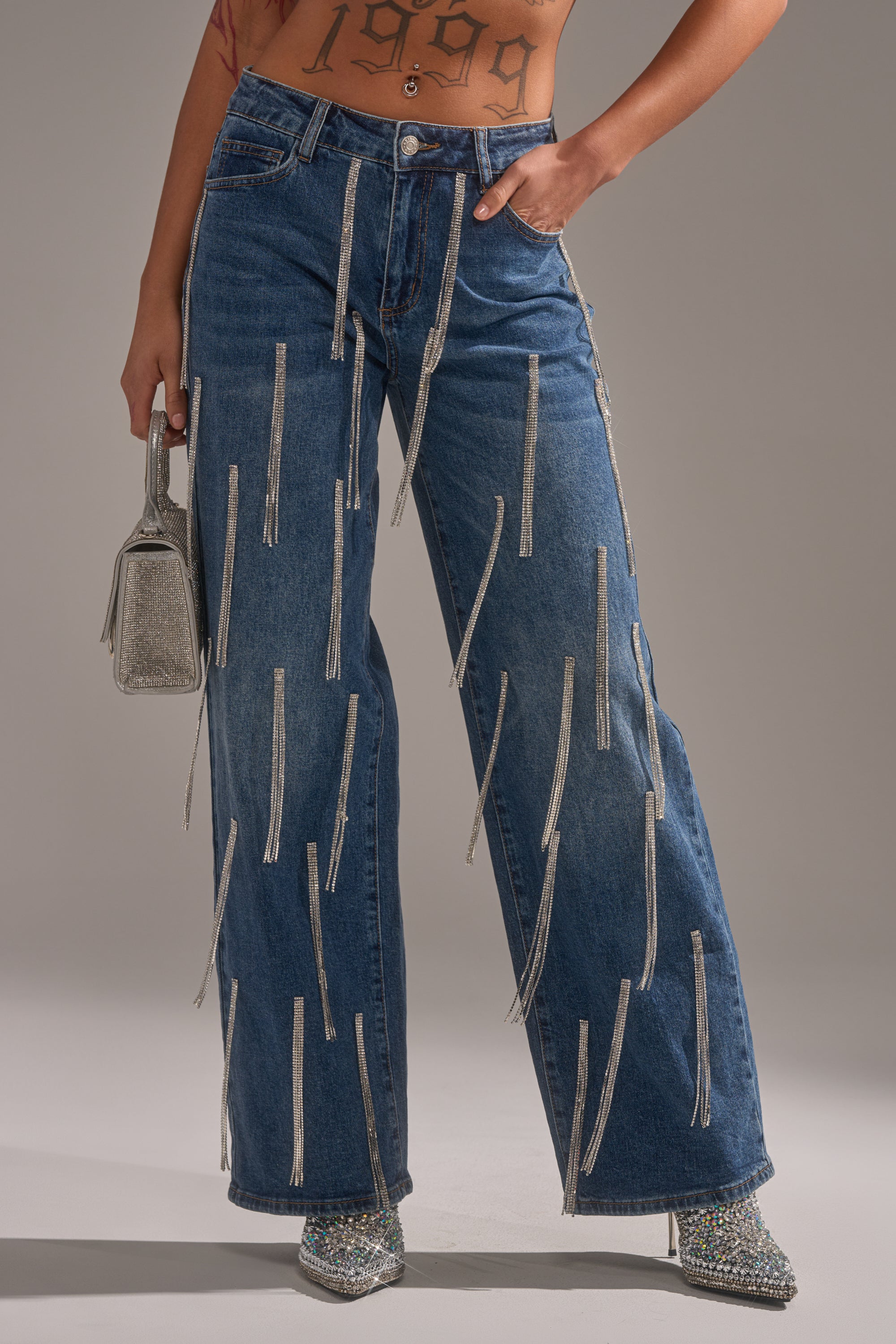 A person wears DRIPPED OUT RELAXED DENIM high-waisted blue jeans with rhinestone chain embellishments, sparkling pointed-toe shoes, holds a glittery silver handbag, and has a "1999" tattoo on their torso.