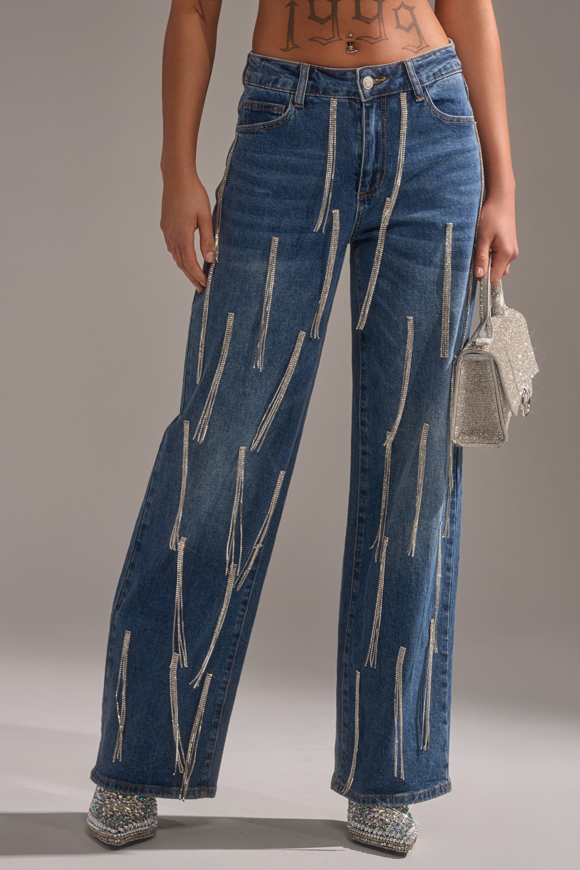 Wearing DRIPPED OUT RELAXED DENIM—high-waisted, wide-leg jeans with rhinestone embellishments—paired with sparkly silver heels and a glittery silver mini handbag. Only the lower torso and legs are shown.