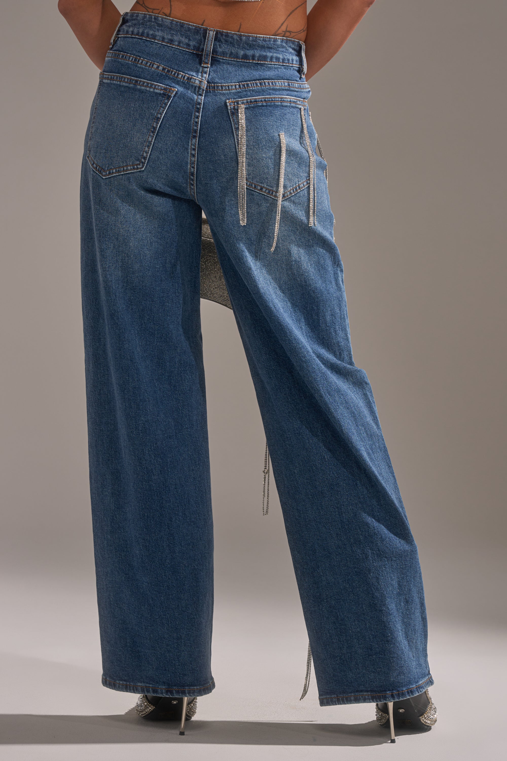 Facing away, a person wears the DRIPPED OUT RELAXED DENIM—high-waisted blue jeans with overlapping belt loops, visible stitching, and a wide-leg fit—paired with heeled shoes against a plain neutral background.