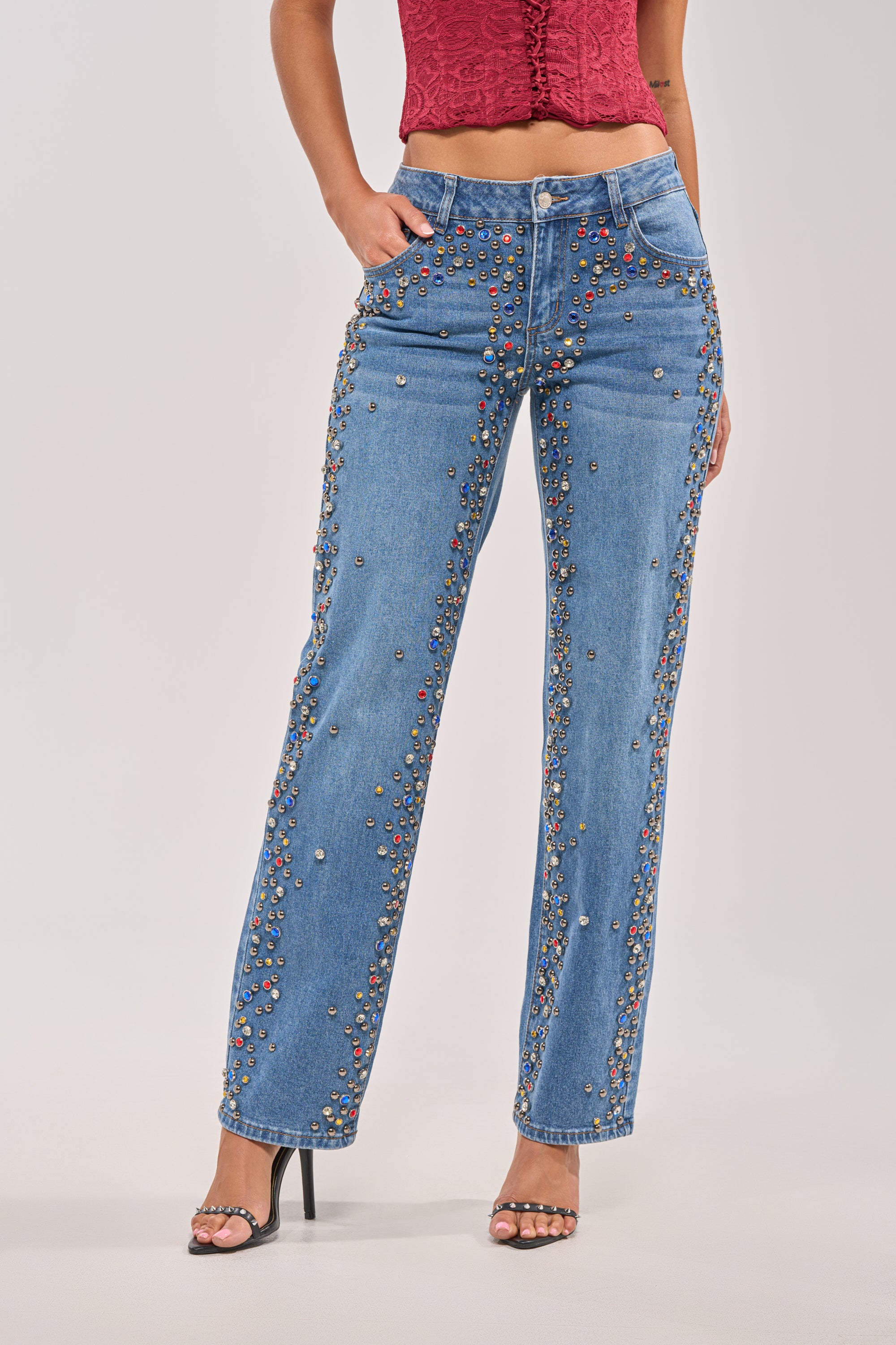 A person wearing the JESSIE RHINESTONE DENIM jeans with a red sleeveless top and black high-heeled sandals stands with one hand in their pocket against a plain background. 0