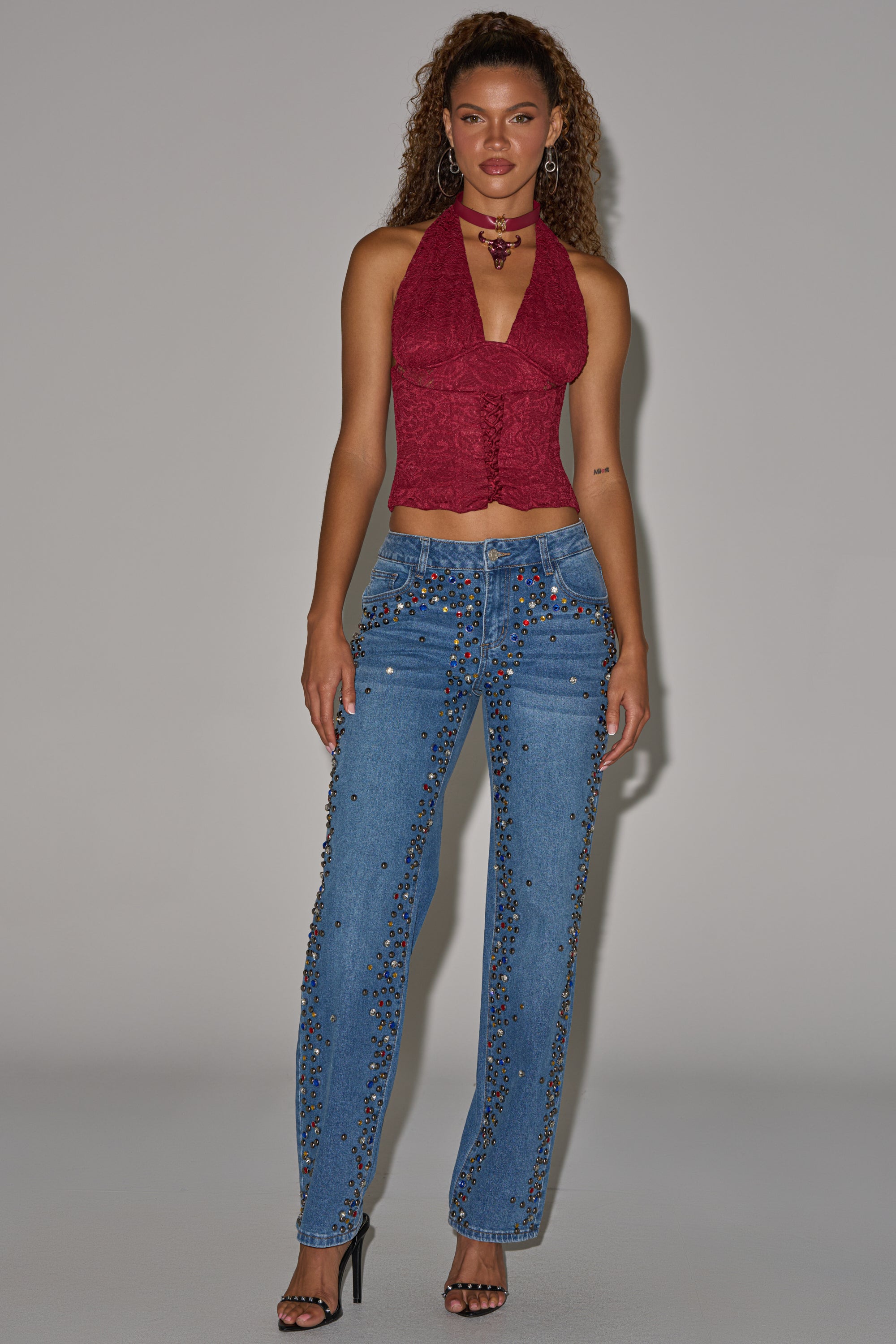 A woman with curly hair poses against a plain backdrop wearing JESSIE RHINESTONE DENIM jeans featuring colorful gem embellishments, a sleeveless red halter top, black heels, and jewelry. 9