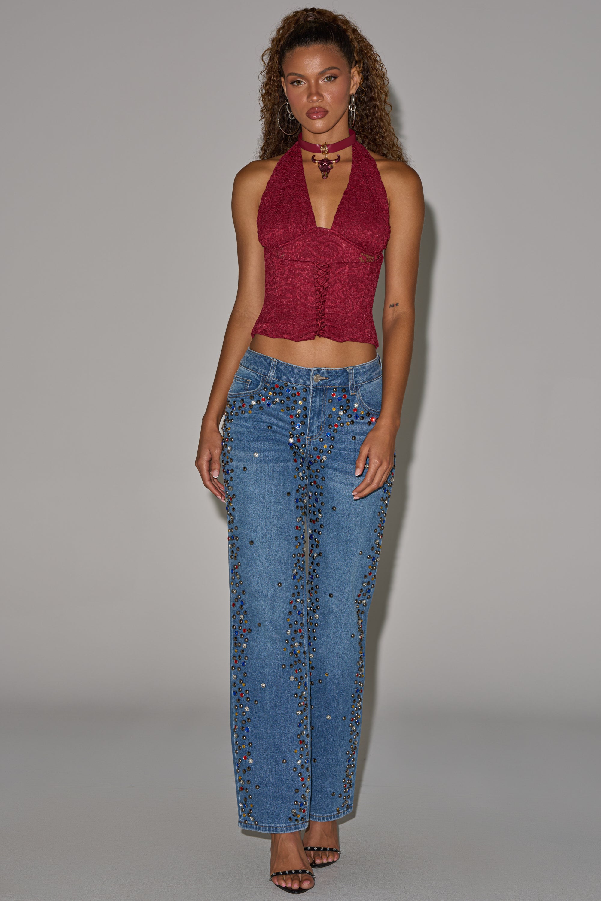 A woman stands against a plain background wearing a sleeveless red halter top, black heels, jewelry, and the JESSIE RHINESTONE DENIM—blue high-waisted dark-wash wide-leg jeans with stud details. 8