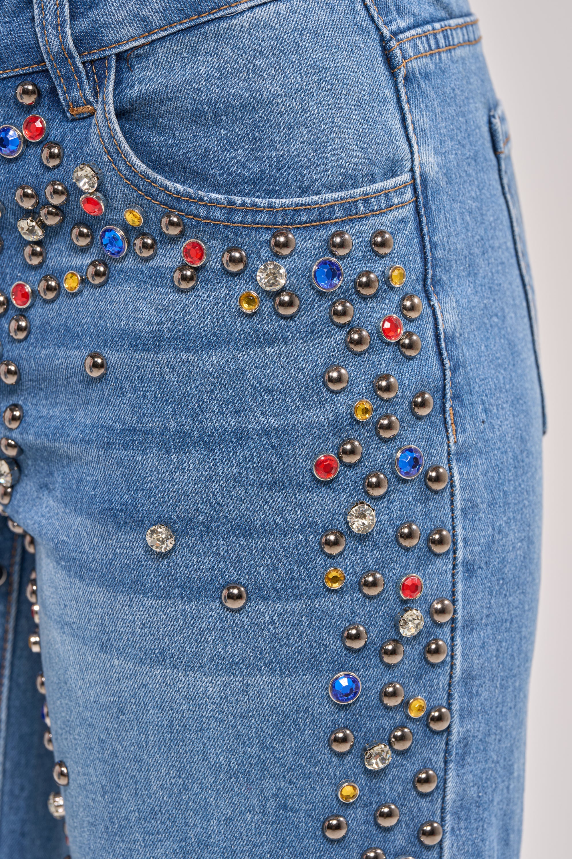Close-up of the JESSIE RHINESTONE DENIM wide-leg jeans featuring multicolored studs and rhinestone embellishments—red, blue, yellow, black, and clear gems clustered on the front pocket and upper thigh. 10