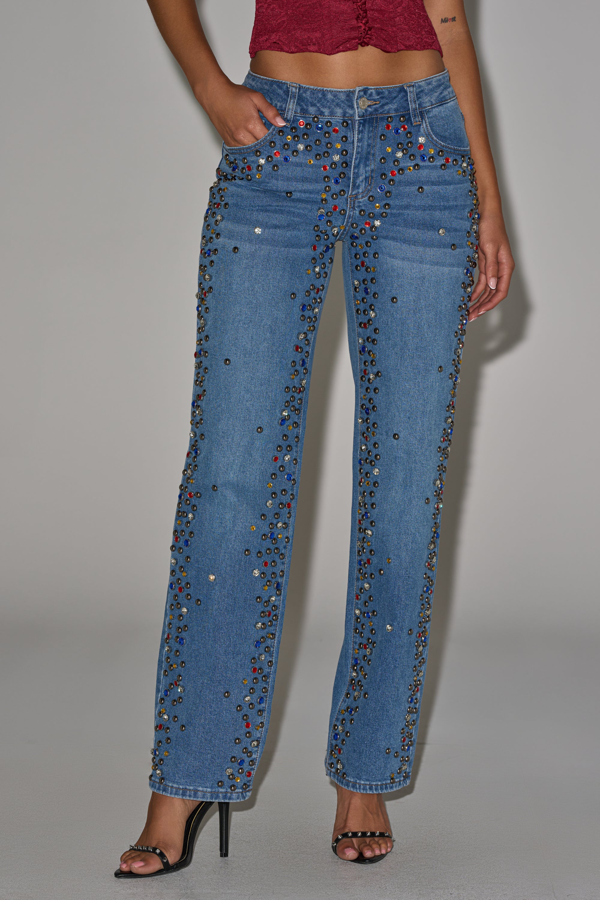 A person wears JESSIE RHINESTONE DENIM wide-leg, dark-wash jeans with colorful studs and a textured red crop top, paired with black high-heeled sandals, standing against a plain background. 1