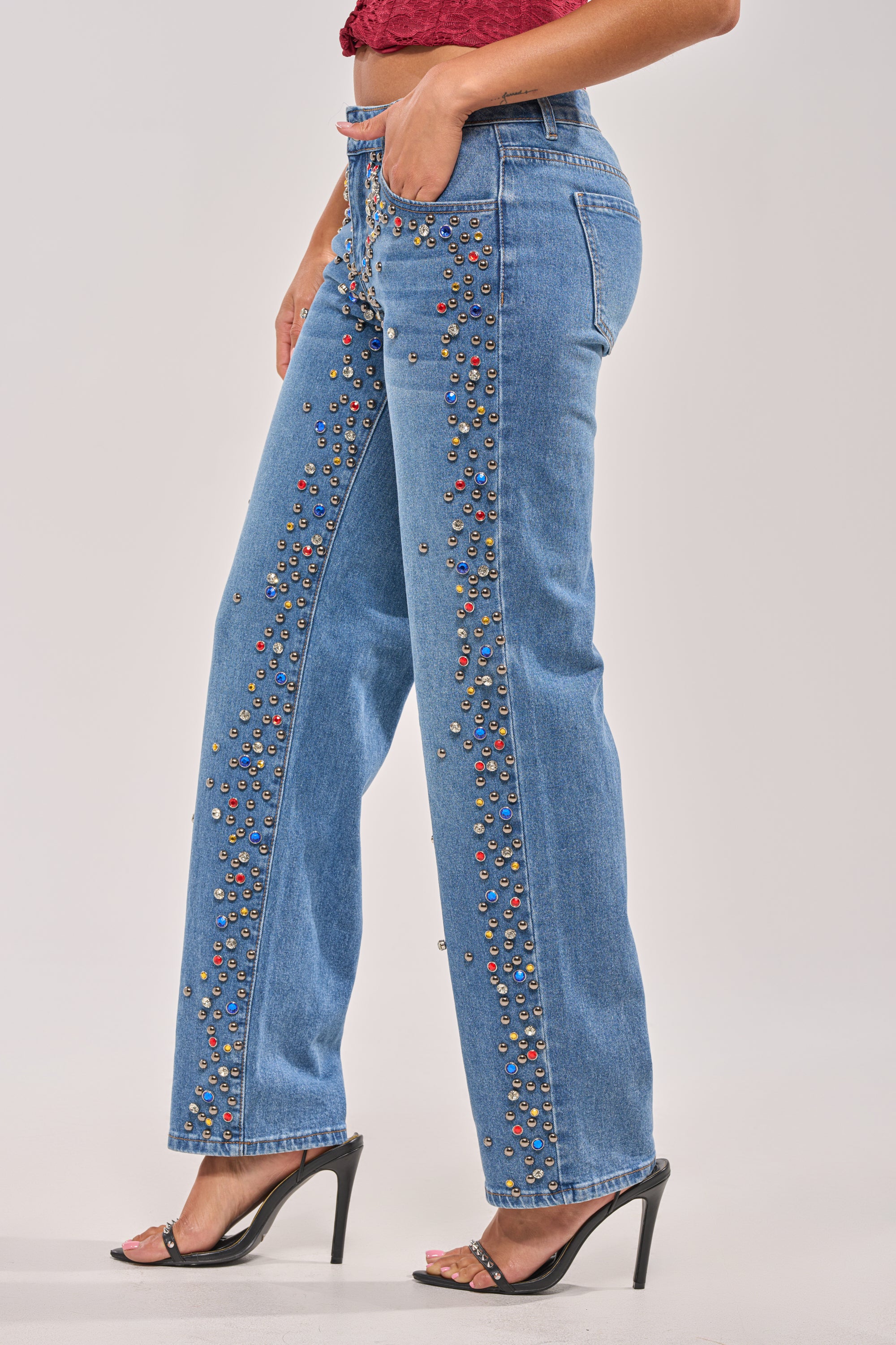 Wearing JESSIE RHINESTONE DENIM wide-leg jeans, a red top, and black high-heeled sandals, a person stands against a plain light background with one hand in their pocket. 3