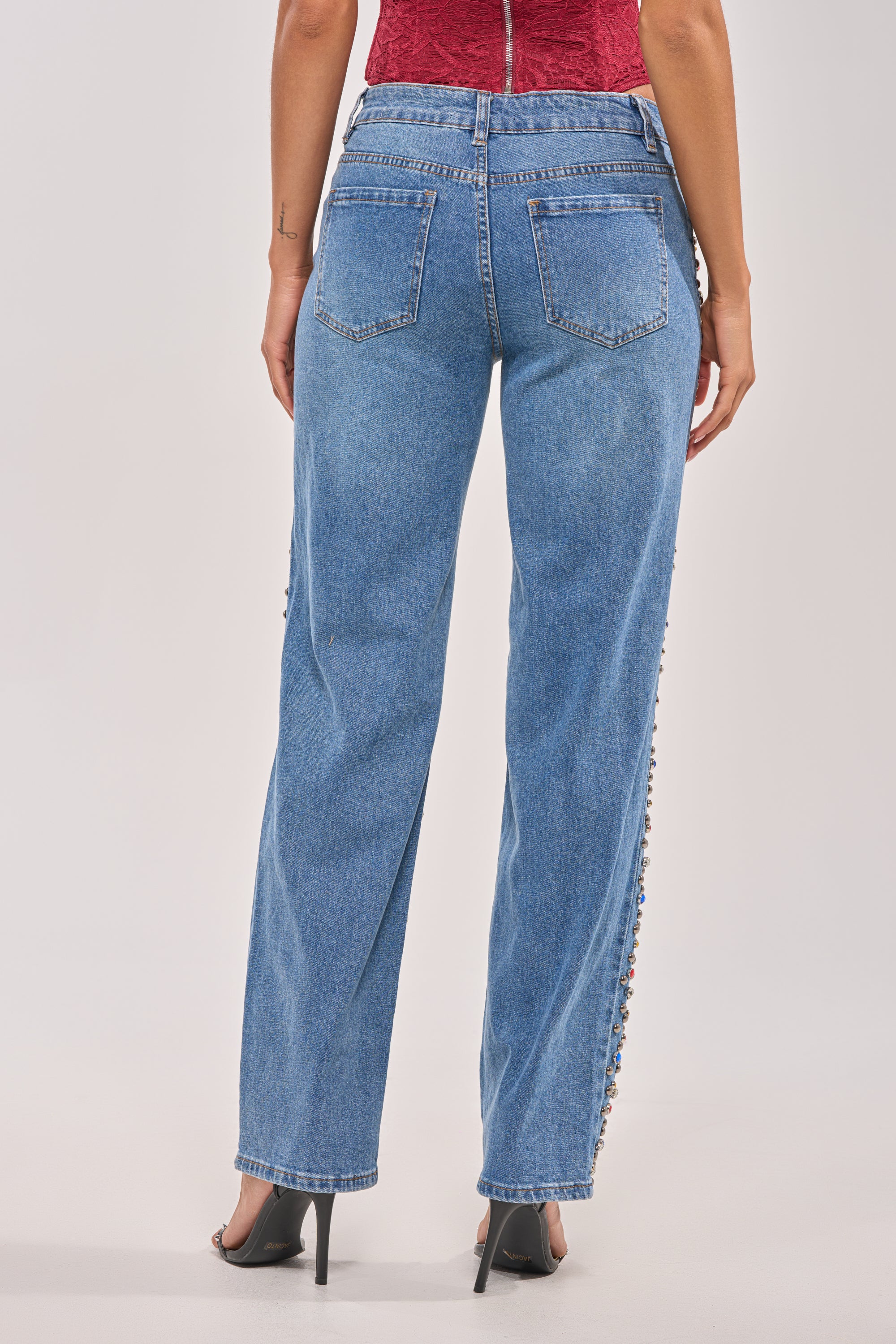 A person stands with their back to the camera wearing JESSIE RHINESTONE DENIM high-waisted, wide-leg blue jeans with back pockets, a sleeveless red top, and black heeled shoes. 4