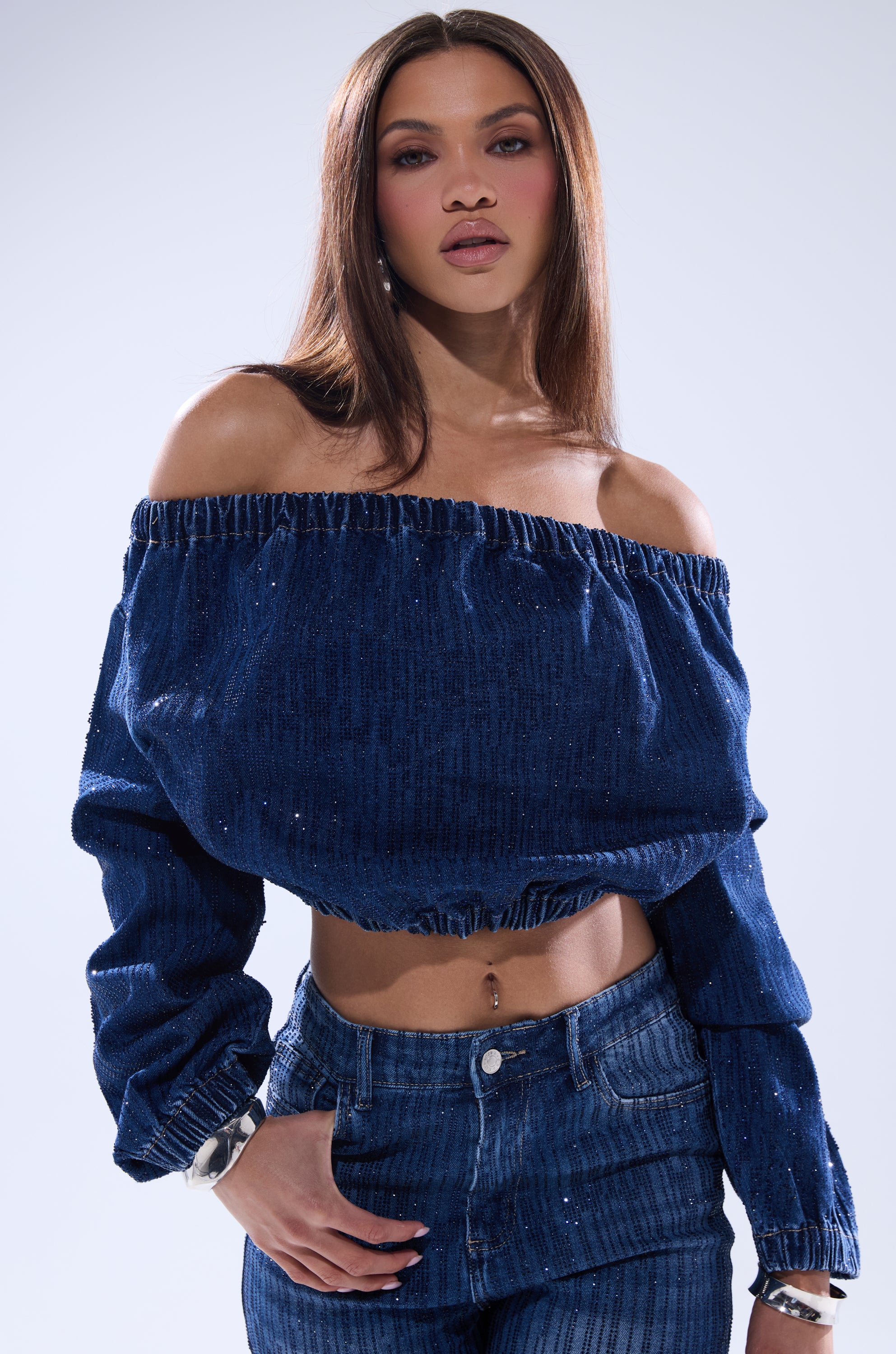 A woman with long straight hair wears the SHINING STAR OFF THE SHOULDER RHINESTONE DENIM SWEATSHIRT and matching high-waisted jeans, standing against a plain light background and confidently gazing at the camera with one hand in her pocket.