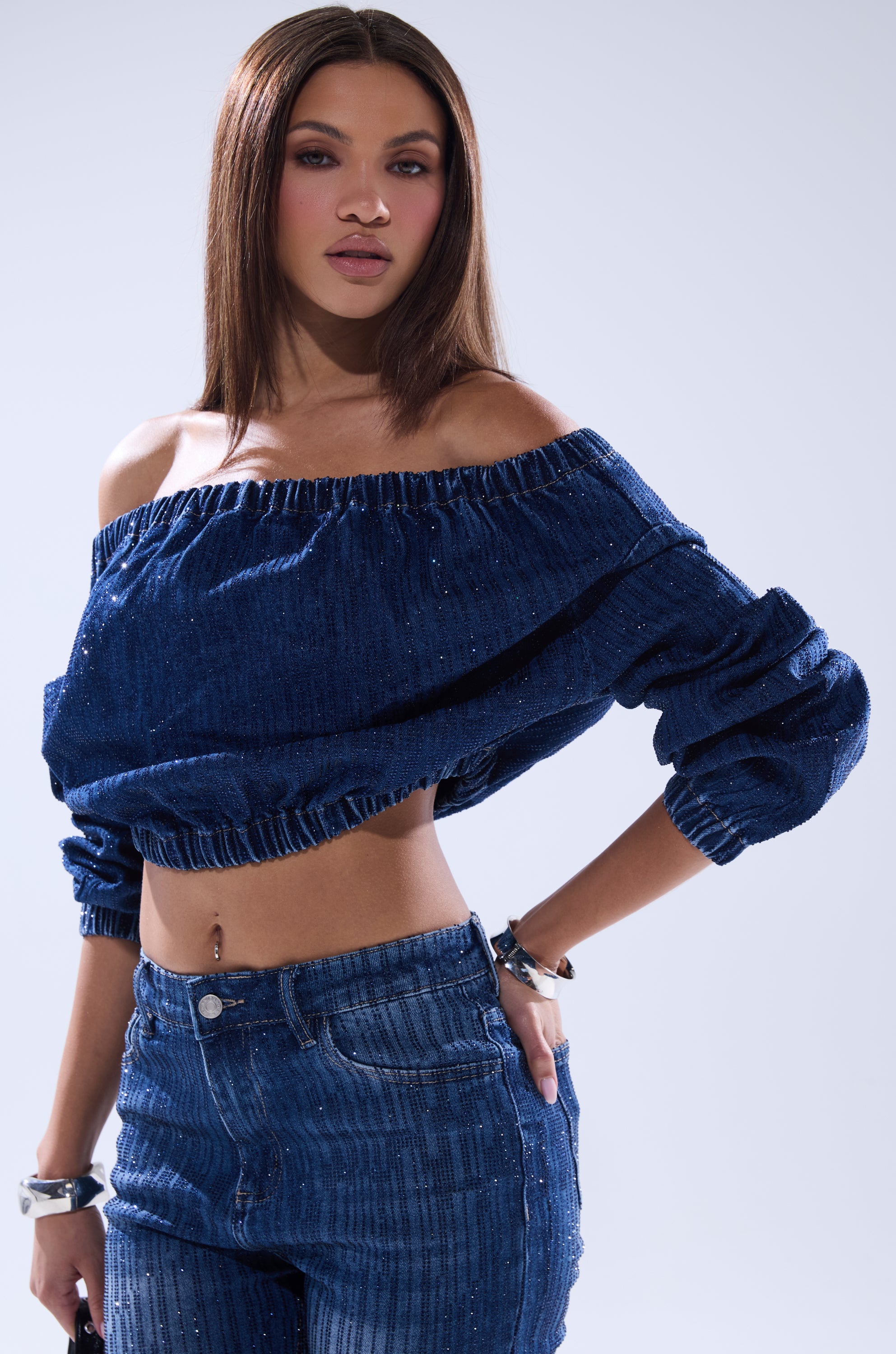 A woman with straight brown hair models the SHINING STAR OFF THE SHOULDER RHINESTONE DENIM SWEATSHIRT and matching pants, posing confidently against a light background to highlight her relaxed, modern look.