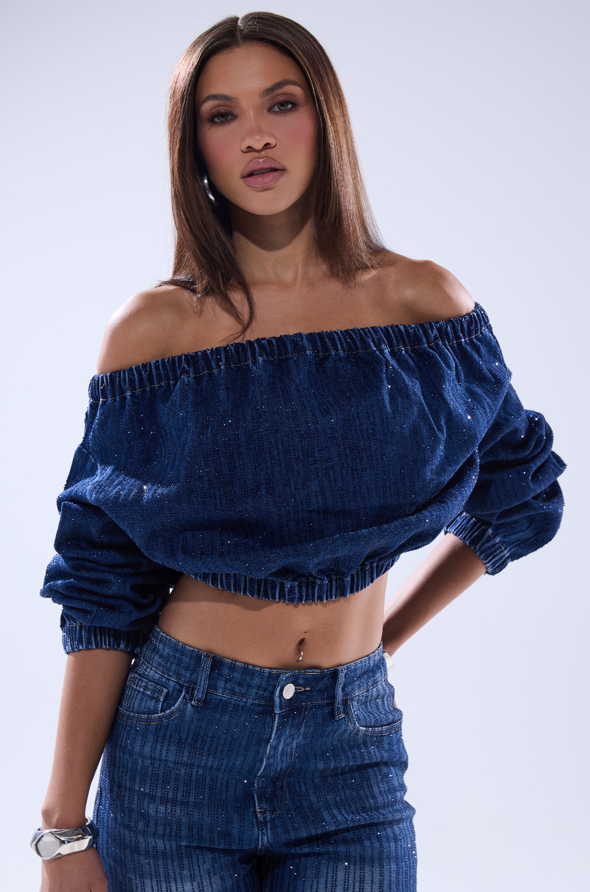 A woman with straight brown hair wears the SHINING STAR OFF THE SHOULDER RHINESTONE DENIM SWEATSHIRT and high-waisted jeans, standing confidently with one arm bent against a plain light background.