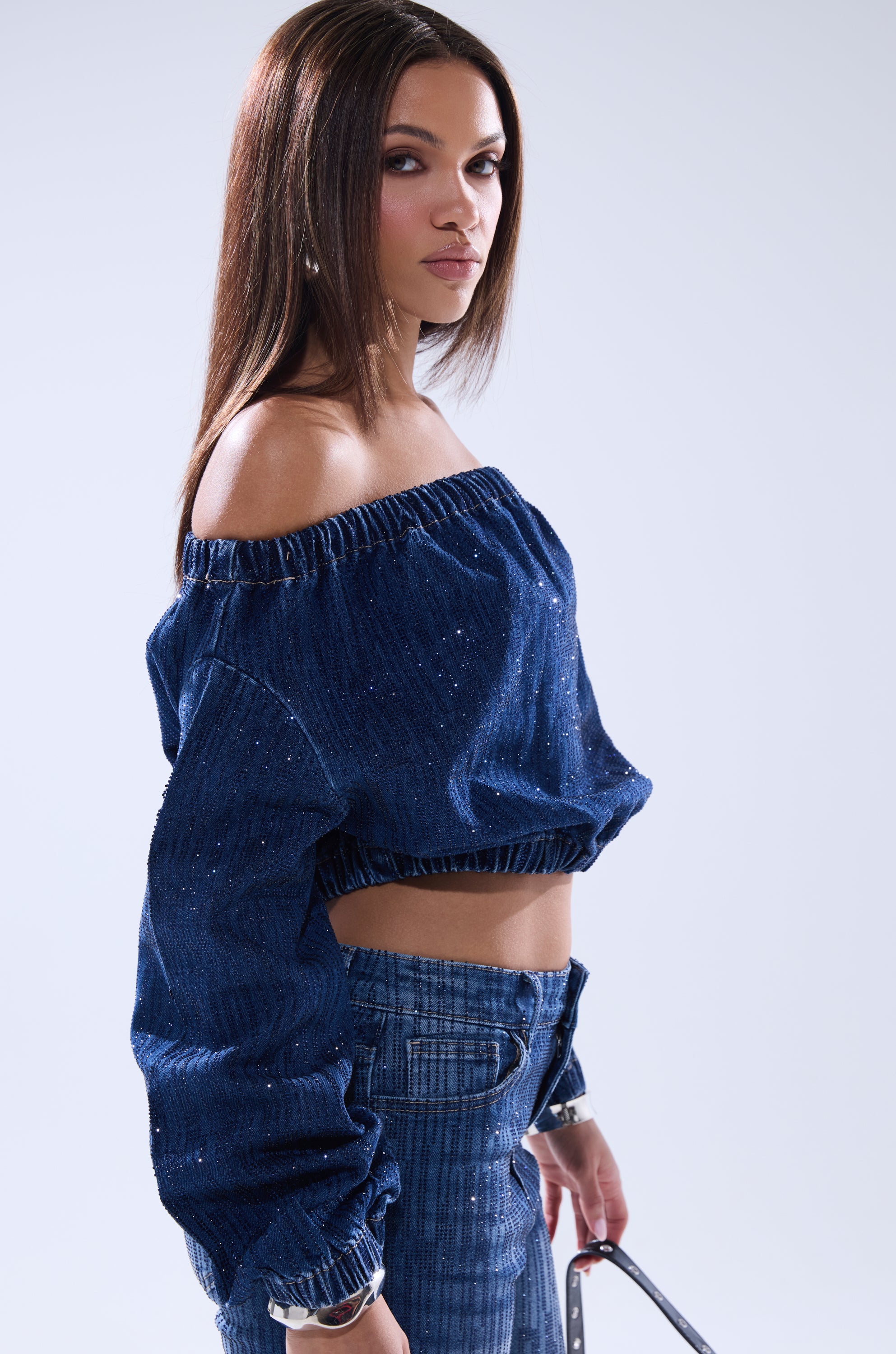 A woman with straight brown hair models the SHINING STAR OFF THE SHOULDER RHINESTONE DENIM SWEATSHIRT and high-waisted denim pants, standing sideways and looking at the camera against a plain light background.