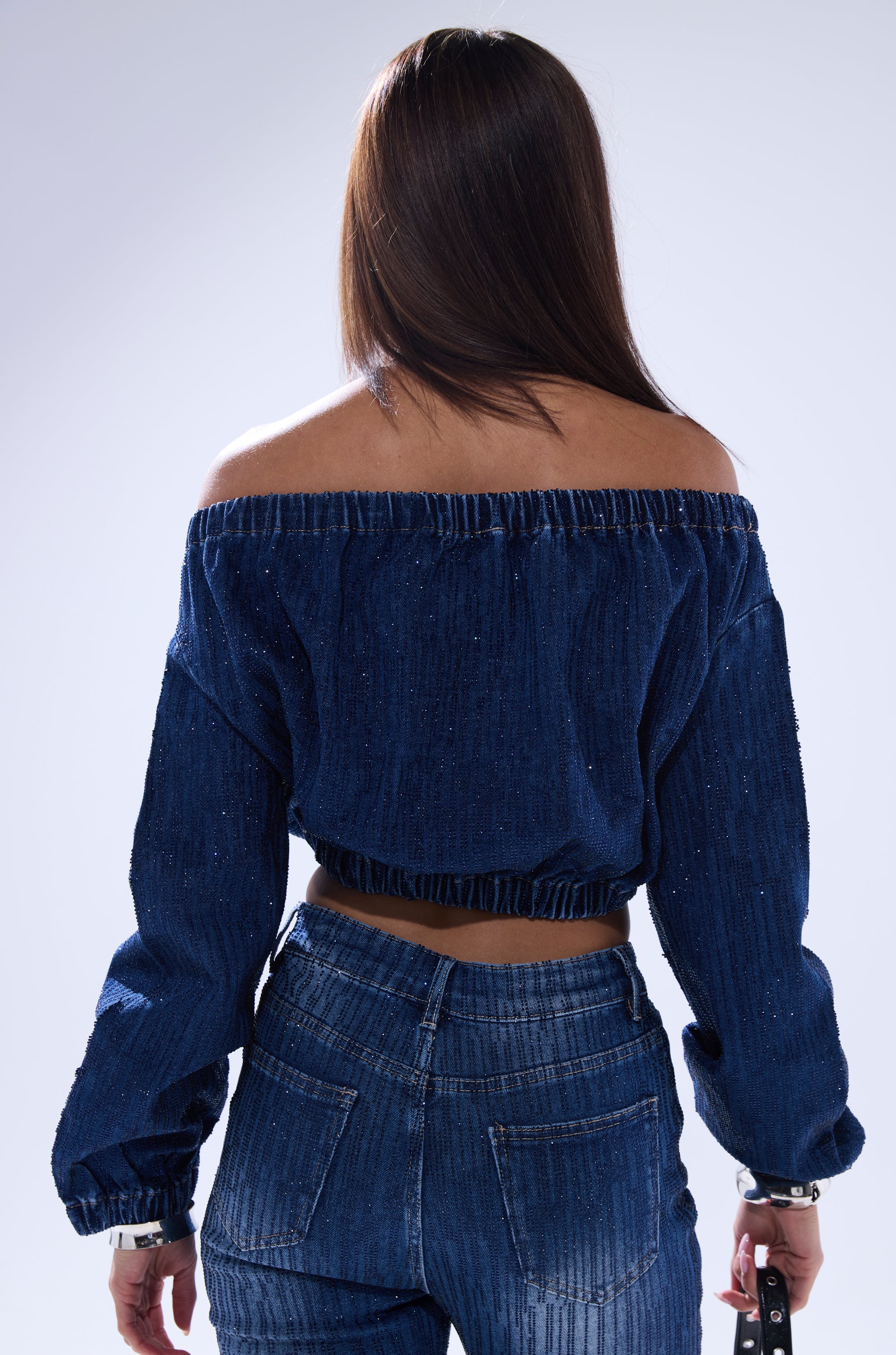 A woman with straight brown hair faces away, wearing the SHINING STAR OFF THE SHOULDER RHINESTONE DENIM SWEATSHIRT and matching high-waisted jeans, holding a small black item in her right hand.