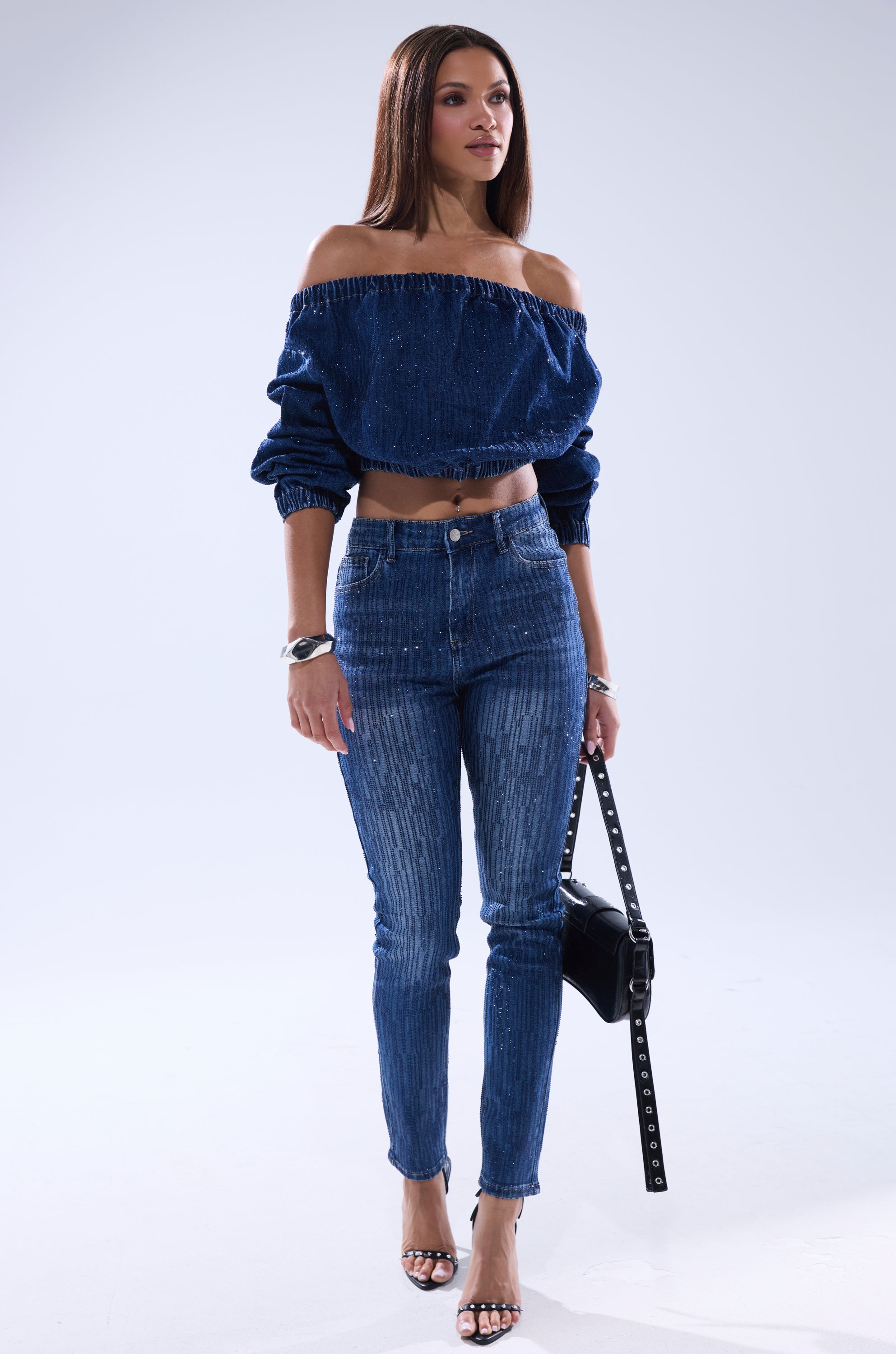 A woman wears the SHINING STAR OFF THE SHOULDER RHINESTONE DENIM SWEATSHIRT with high-waisted jeans, black handbag, and heeled sandals. Her straight brown hair frames her face as she poses against a plain light background.
