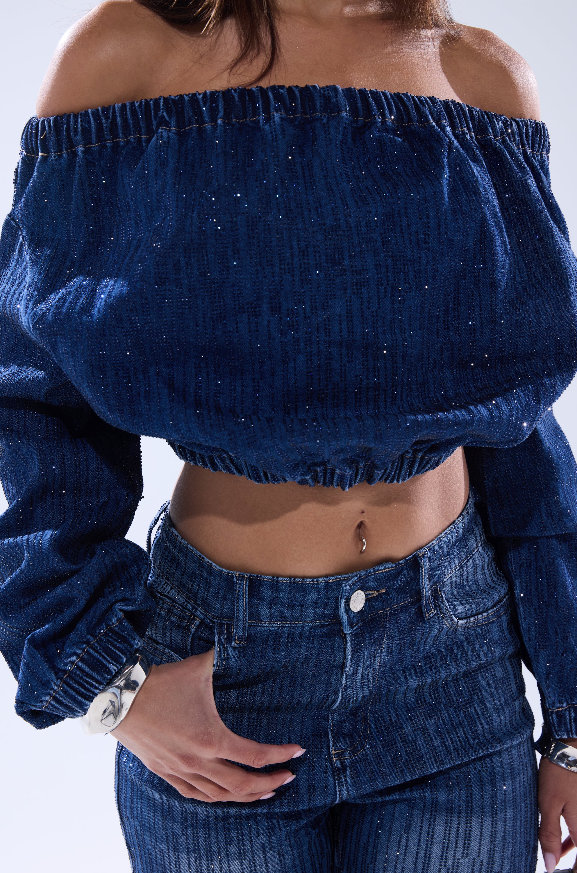 Wearing the SHINING STAR OFF THE SHOULDER RHINESTONE DENIM SWEATSHIRT with matching high-waisted jeans, this look features a sparkling off-shoulder top that reveals the midriff and highlights a navel piercing.