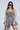 A woman with long dark hair and sunglasses poses against a light blue background wearing the VIVIAN DENIM MINI DRESS. The gray mini dress features a corset bodice, flared skirt, and seam details for an edgy touch. She carries a black bag to complete her chic look.