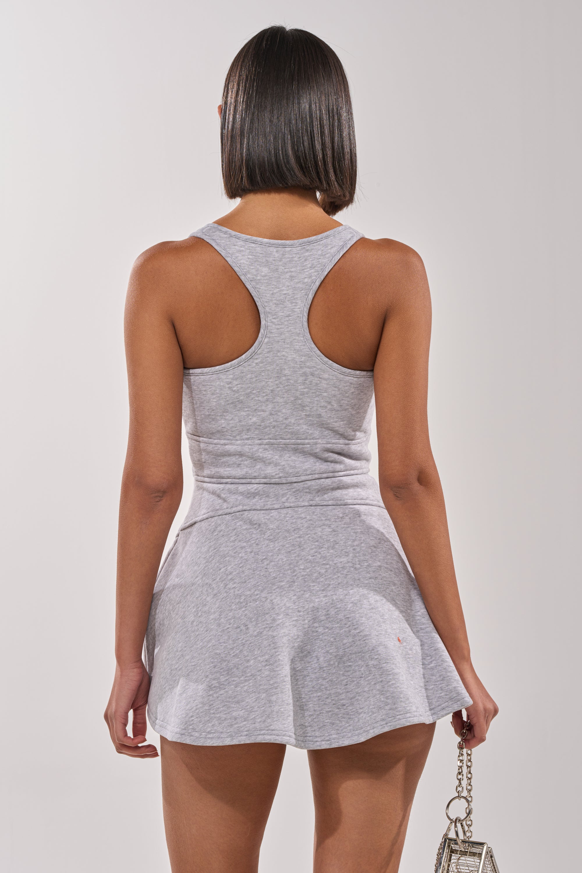 A person with short, straight hair is shown from the back in the MATCH POINT MINI DRESS IN HEATHER GREY, featuring a racerback cut, paired with a small handbag with a metallic chain strap against a plain light background. 3