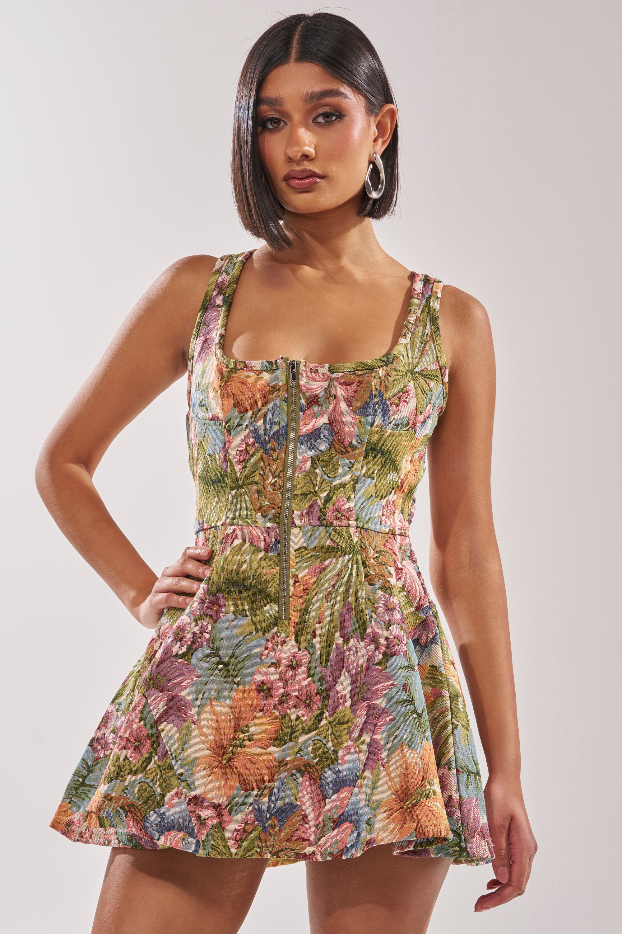 A woman with straight dark hair wears the MATCH POINT MINI DRESS IN BROCADE, a sleeveless jacquard print style with a front zipper. She poses with one hand on her hip against a plain light background. 1