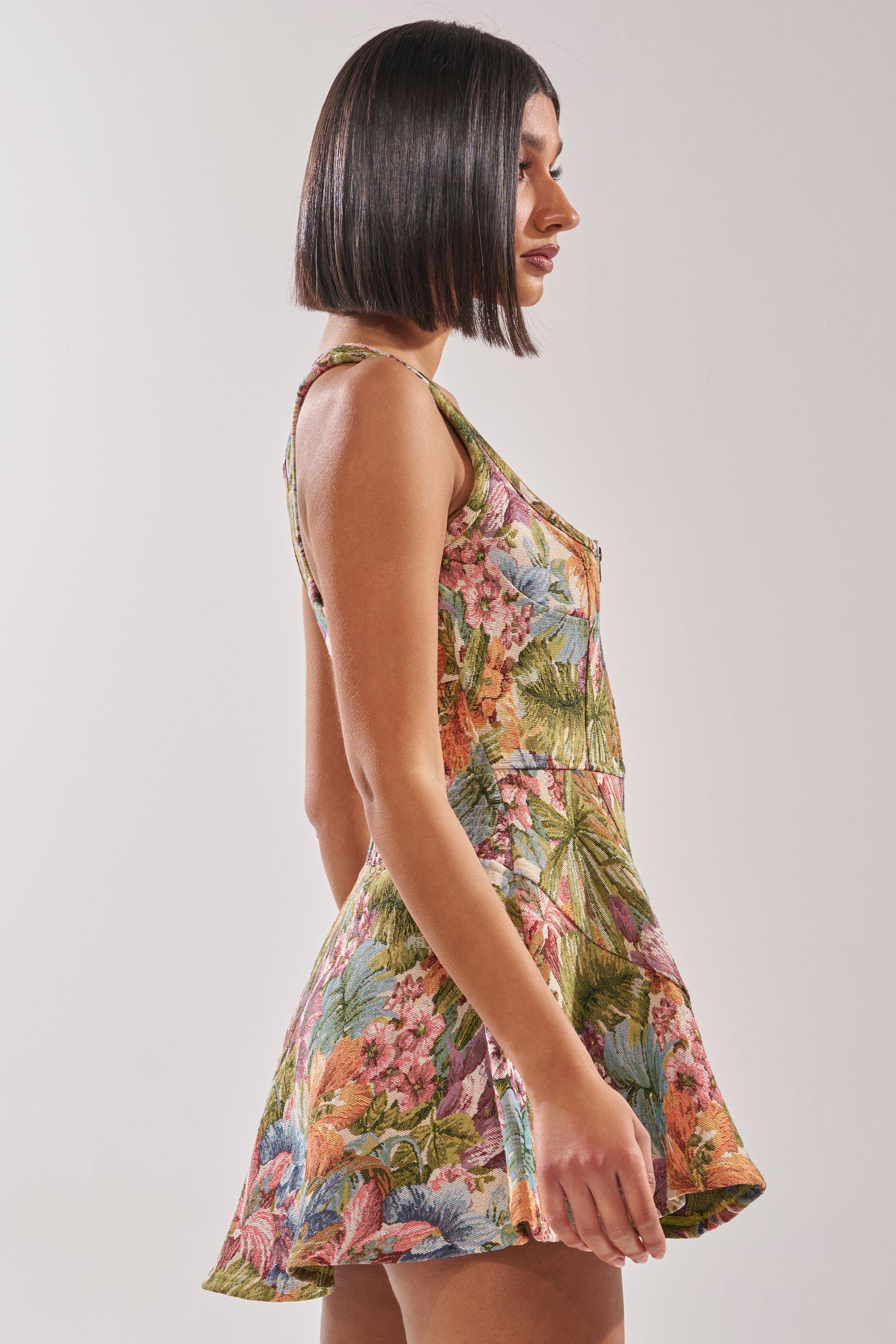 A woman with straight, dark bob-length hair stands in profile, modeling the MATCH POINT MINI DRESS IN BROCADE—a sleeveless skater style with a flared skirt—against a plain light background. 3