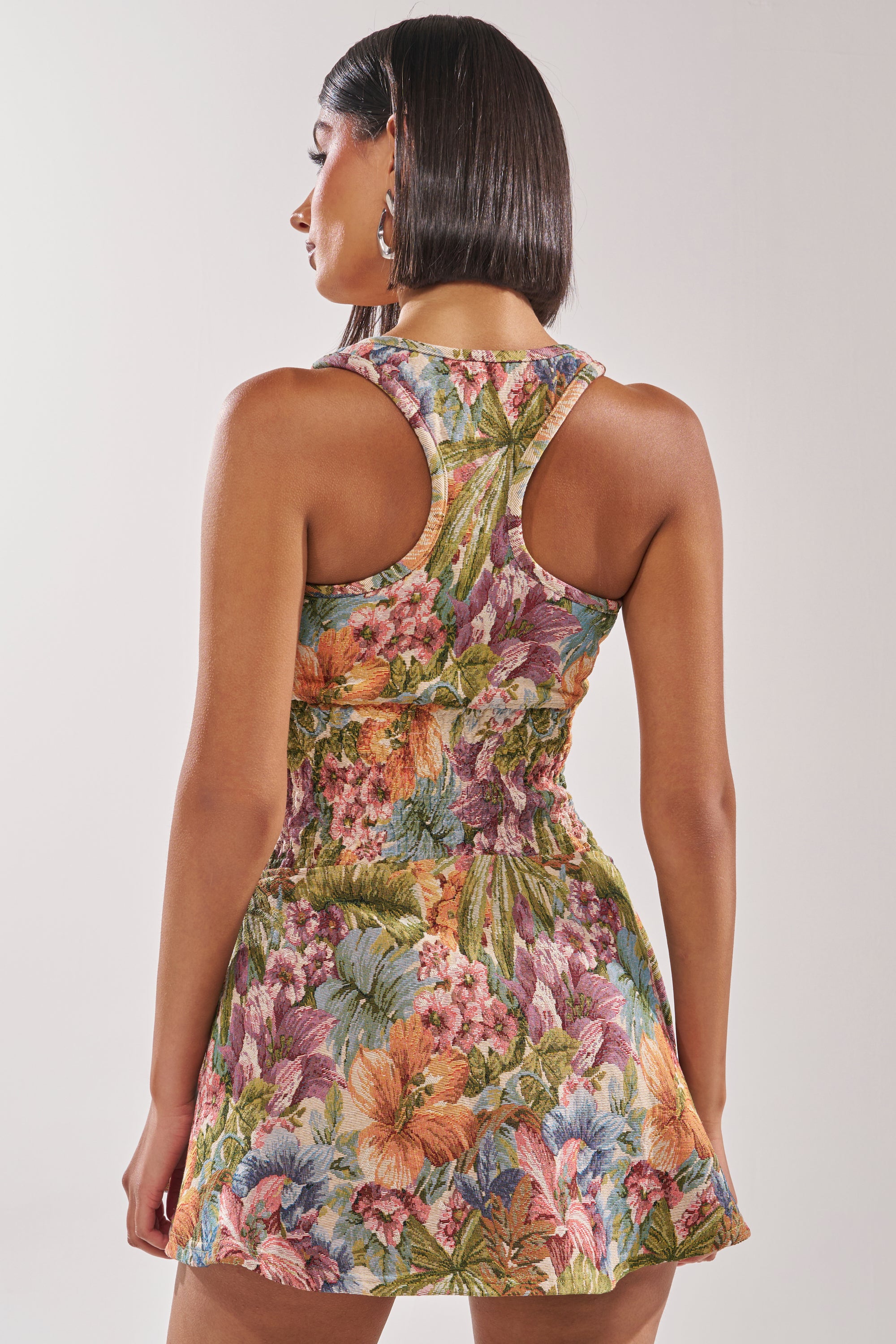 A woman with straight dark hair models the MATCH POINT MINI DRESS IN BROCADE, a sleeveless, racerback skater dress with a colorful floral pattern. She faces away, showcasing the dress's unique back design against a plain background. 4