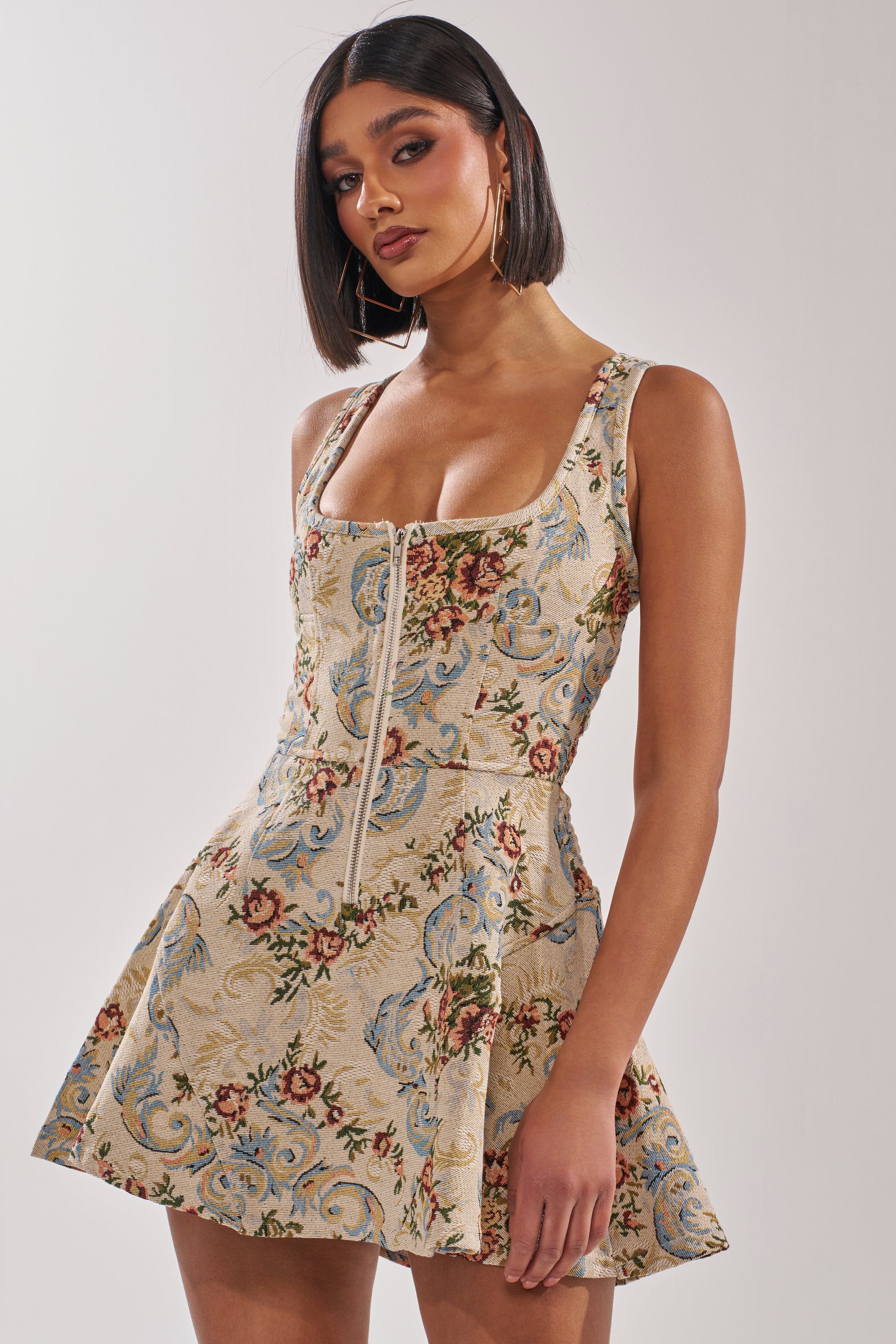 Woman wearing the MATCH POINT MINI DRESS IN LIGHT BROCADE, a sleeveless, square-neck mini dress with floral jacquard fabric, fitted bodice, and flared skirt, posing against a plain background. 1