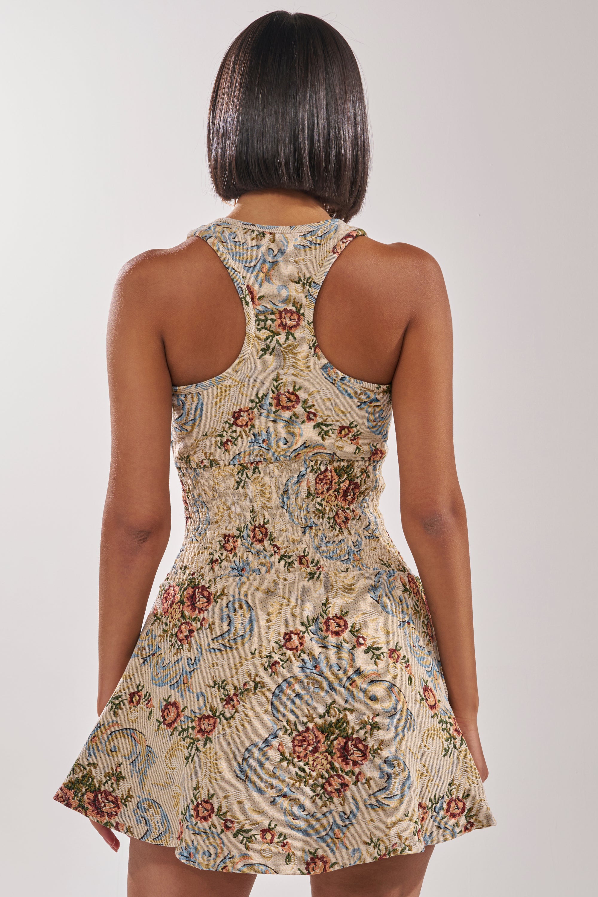 A woman with straight, shoulder-length hair is seen from behind wearing the MATCH POINT MINI DRESS IN LIGHT BROCADE—a sleeveless, vintage-inspired dress with a beige base, blue floral pattern, fitted bodice, and flared skirt. 3
