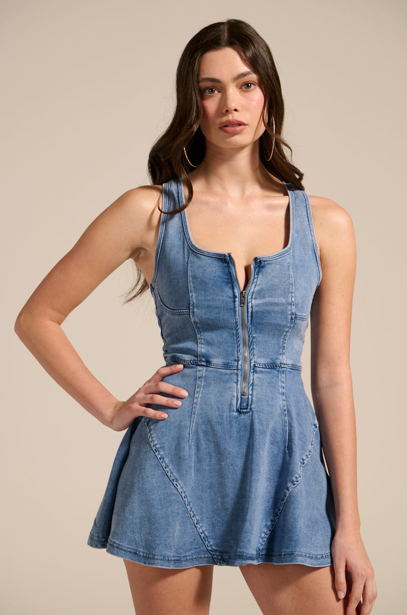In a punk-inspired look, a woman wears the VIVIAN DENIM MINI DRESS with a front zipper. With her hand on her hip and long hair cascading down, she confidently gazes at the camera against a plain backdrop.