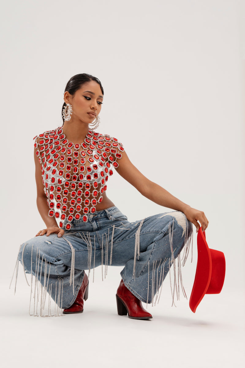 Against a neutral background, someone models a shimmering red and silver rhinestone top paired with DRIPPING IN JEWELS RELAXED FIT DENIM JEANS with fringe and red cowboy boots. They're accessorized with a red cowboy hat and large hoop earrings.