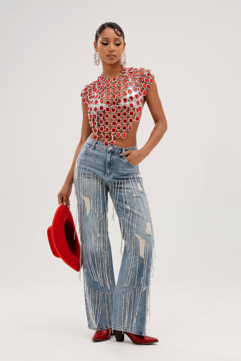 A person confidently wears an open-patterned red top with silver accents and Dripping In Jewels Relaxed Fit Denim Jeans adorned with rhinestones. They hold a red wide-brimmed hat, wear red ankle boots, accessorize with large hoop earrings, and have their hair pulled back.