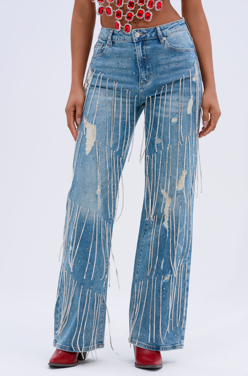 Someone is wearing DRIPPING IN JEWELS RELAXED FIT DENIM JEANS with rhinestone embellishments, dangling beaded fringes, and patches. These blue distressed jeans pair perfectly with a red and white patterned top and red boots against a plain white background.