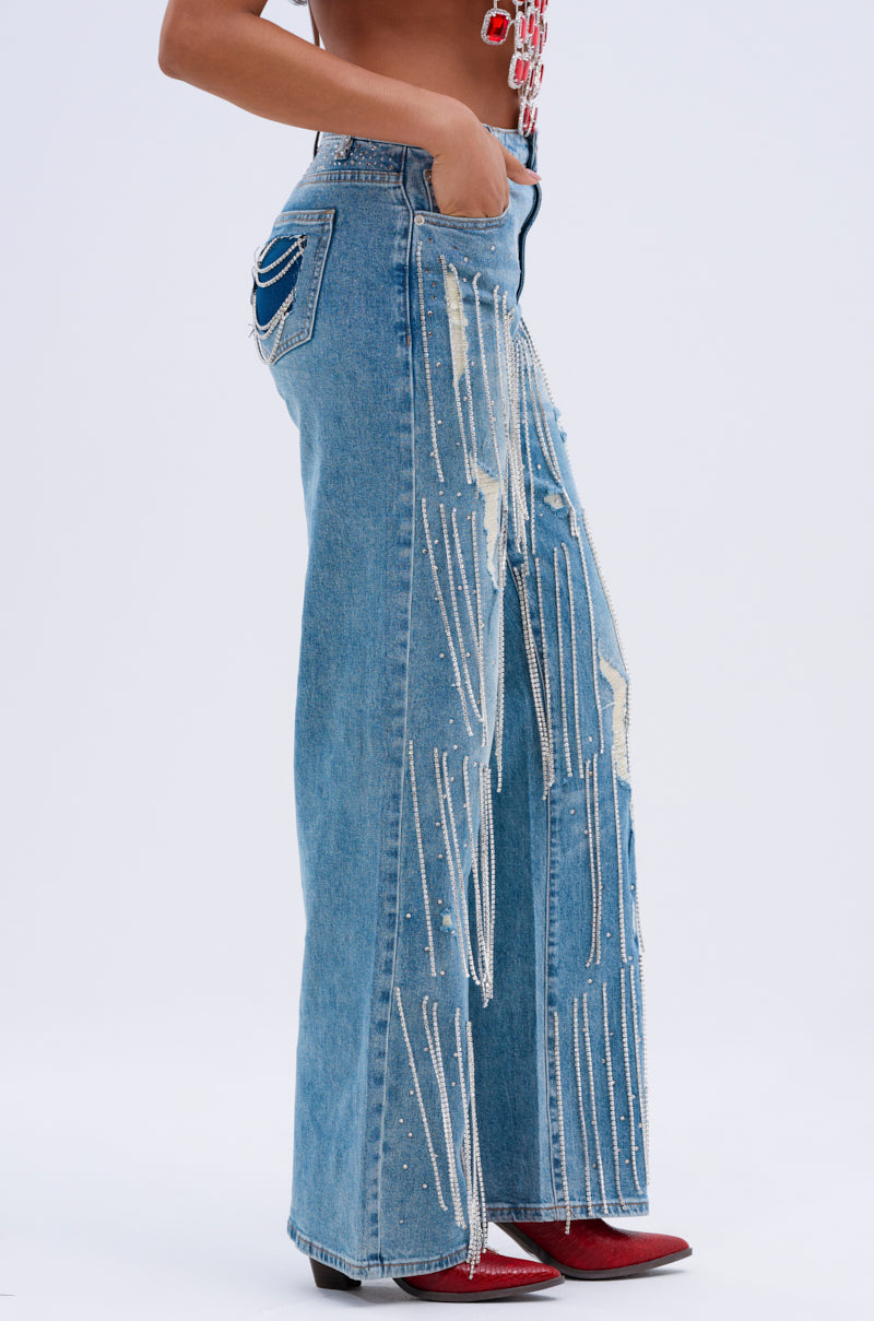 A person in DRIPPING IN JEWELS RELAXED FIT DENIM JEANS, featuring wide-leg cut with distressed details, rhinestone embellishments, frayed design, and hanging chains stands sideways. They're wearing red shoes with a black heel against a plain white background.