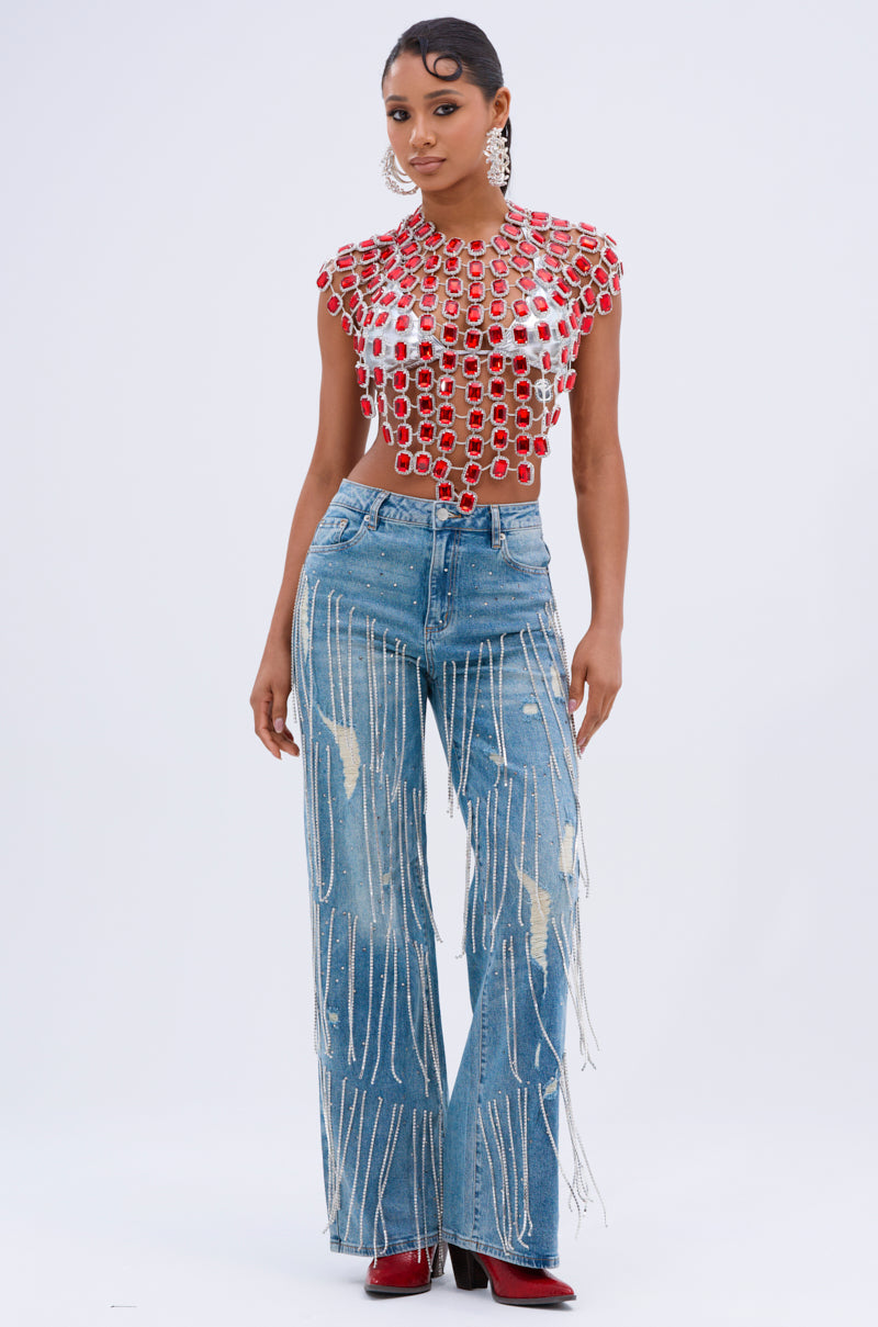 A person poses in a stylish outfit featuring a red top with circular embellishments and DRIPPING IN JEWELS RELAXED FIT DENIM JEANS with white splatter patterns, paired with red boots and long earrings against a plain white background.