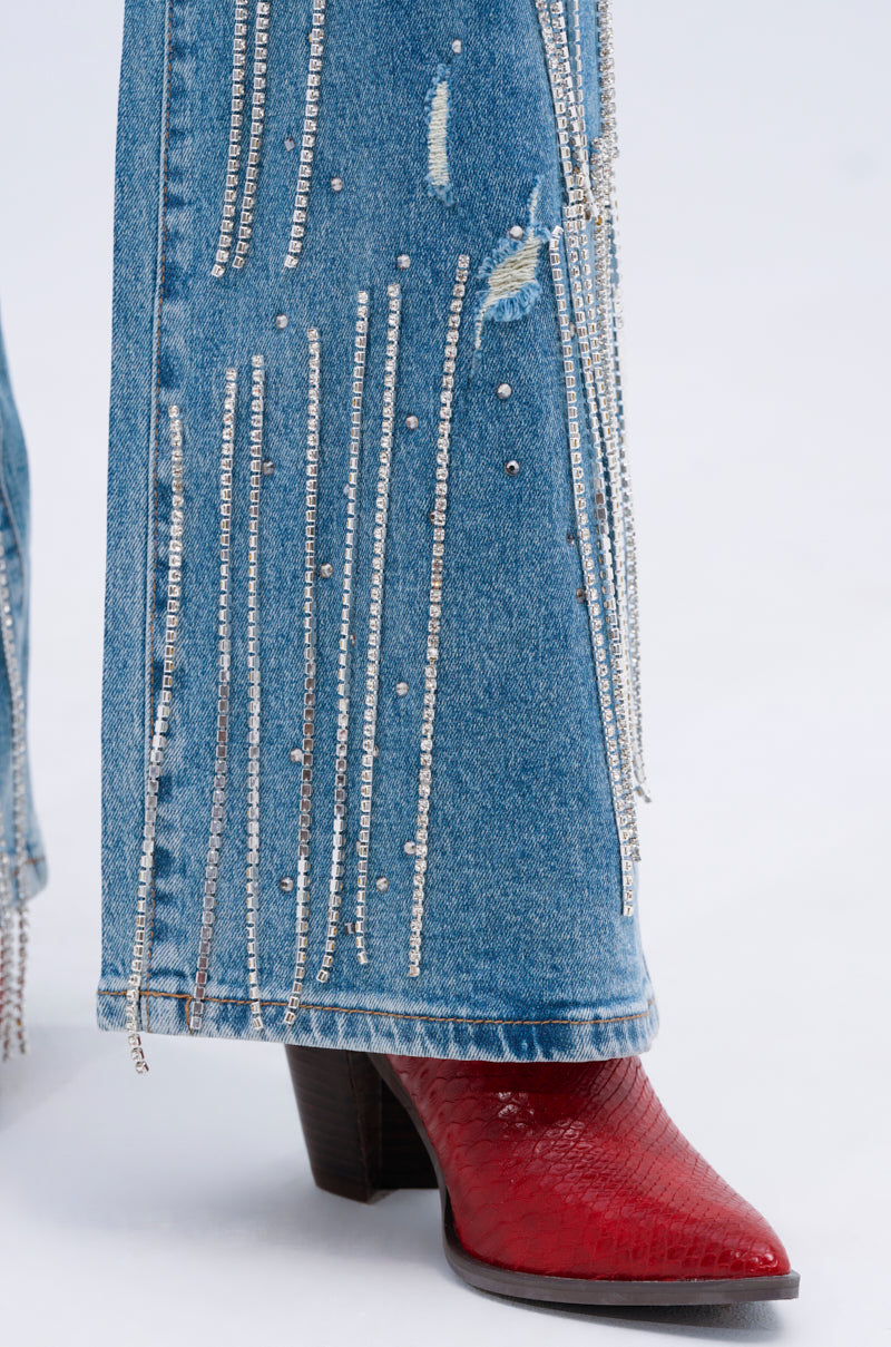 DRIPPING IN JEWELS RELAXED FIT DENIM JEANS feature rhinestone-adorned vertical lines and distressed accents. They elegantly drape over a pointed red snakeskin-pattern boot with a chunky heel, all set against a plain white backdrop.