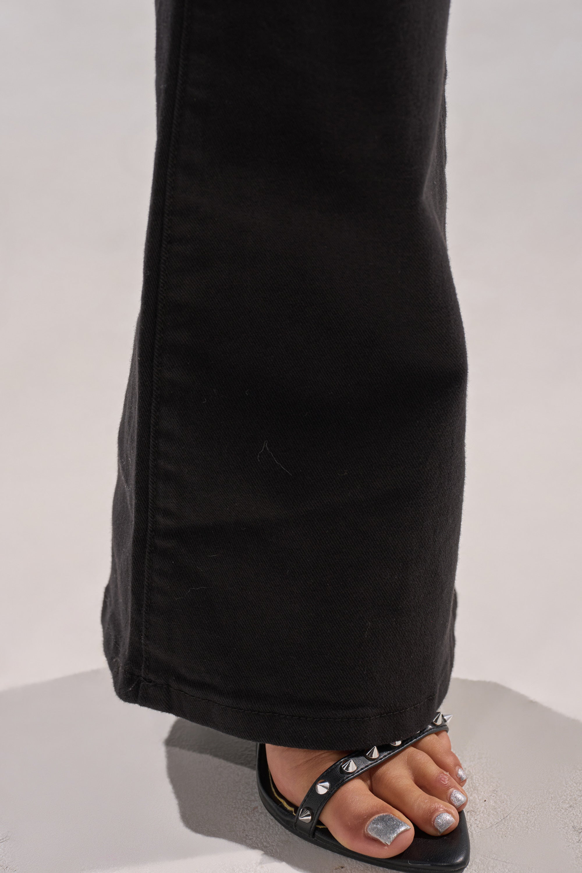 A close-up of a person’s foot in black studded sandals with silver-painted toenails, partially covered by the hem of LOW LIFE LACE-UP JEANS in bootcut black denim, standing on a light-colored surface.