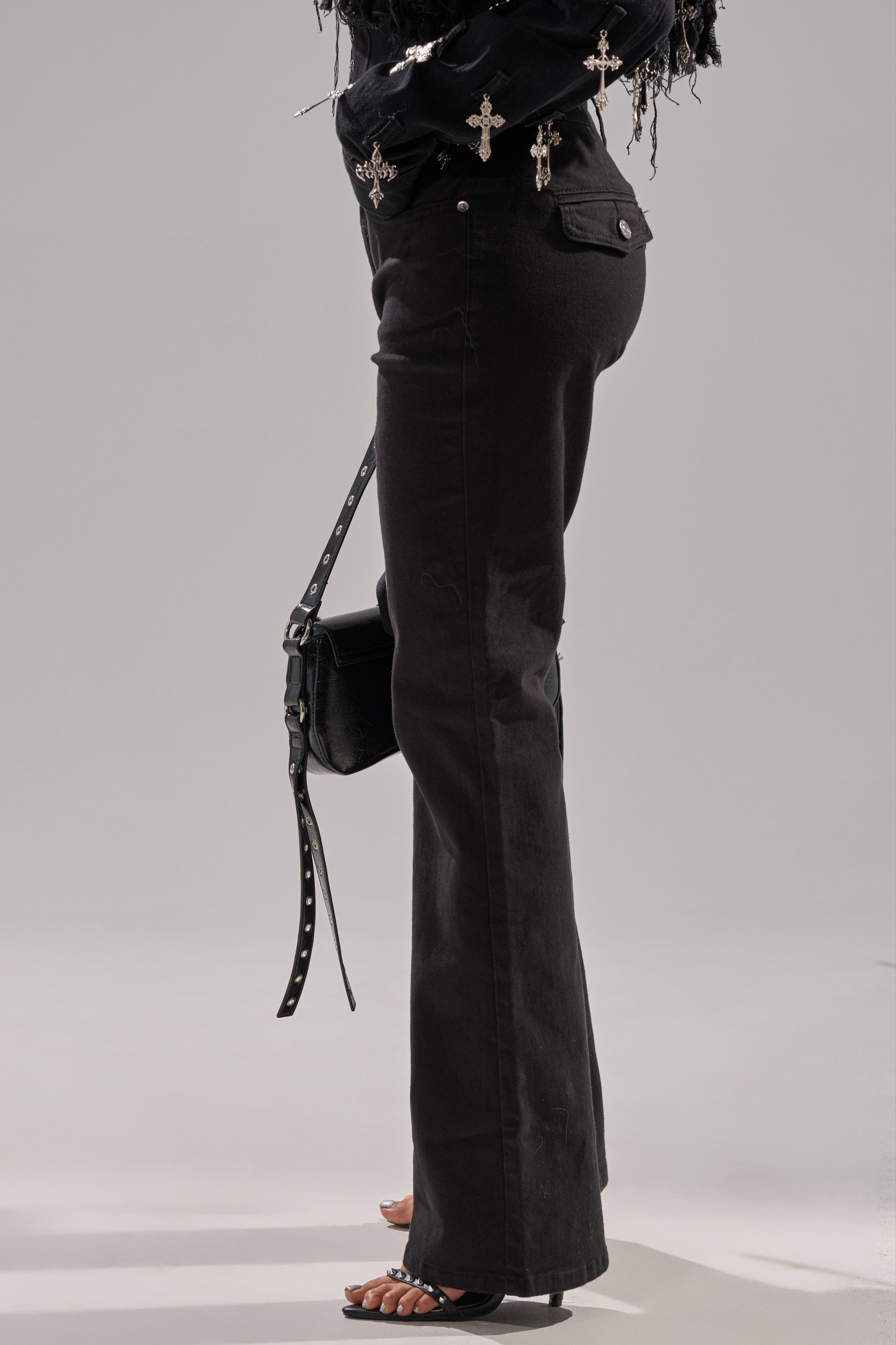A person stands against a plain light gray background wearing LOW LIFE LACE-UP JEANS, a black long-sleeve top with cross-shaped embellishments, open-toe heels, and holds a black purse with a studded strap.