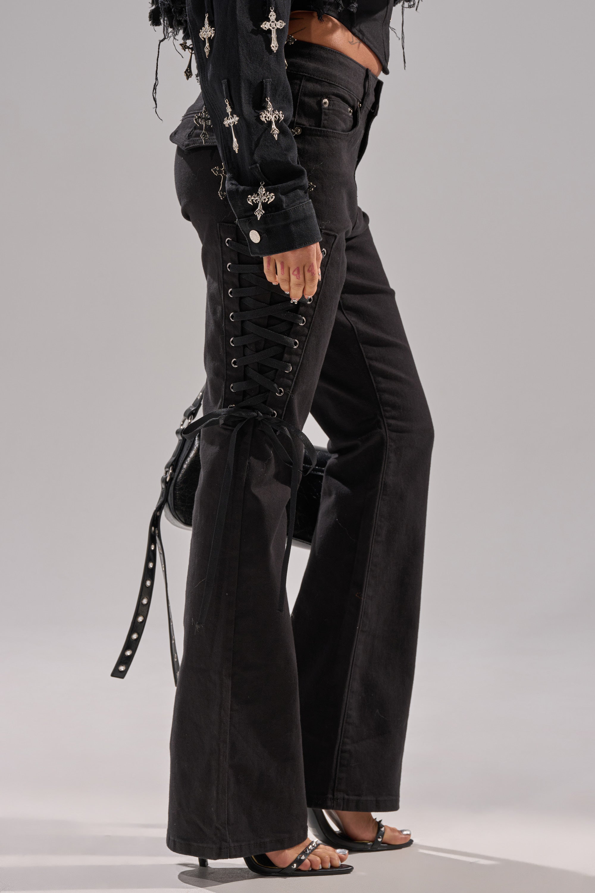 A person wearing LOW LIFE LACE-UP JEANS—black, lace-up, flared-leg denim—paired with a black cross-adorned top, a studded-strap black bag, and open-toe heels.