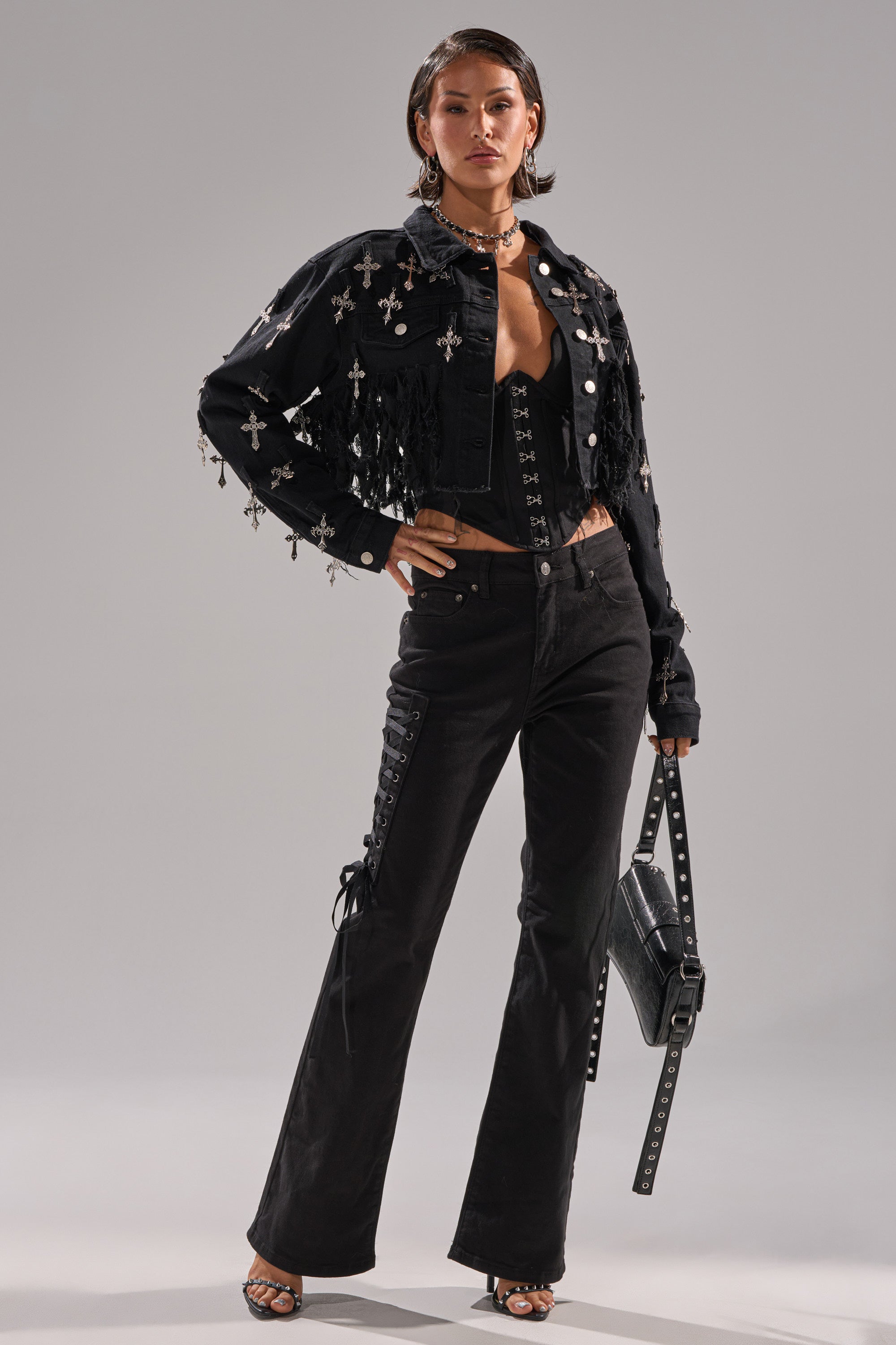 A woman stands confidently in LOW LIFE LACE-UP JEANS, a black fringed denim jacket with silver cross embellishments, a black crop top, heeled sandals, and a studded handbag.