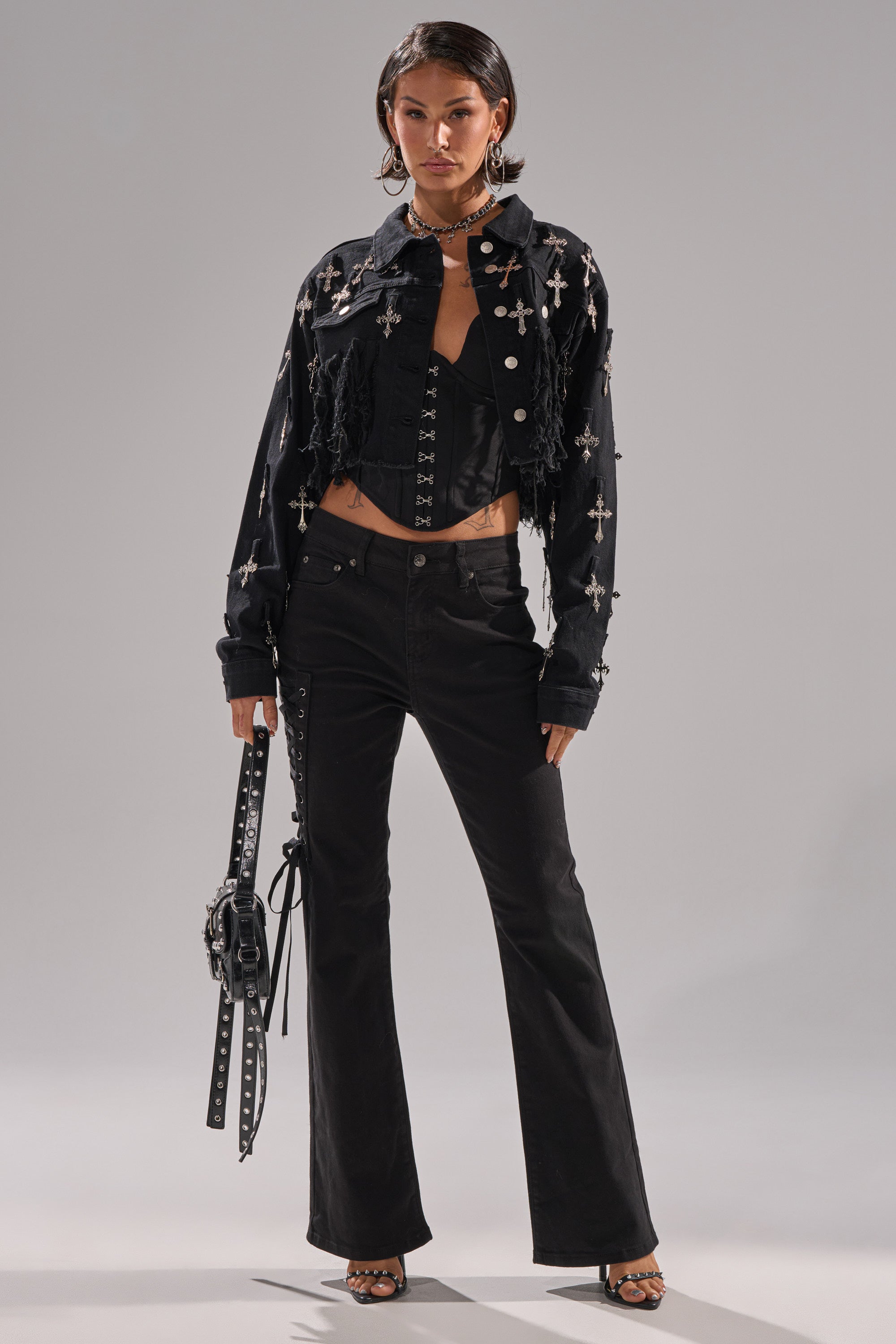 A woman stands confidently in LOW LIFE LACE-UP JEANS with a bootcut flare, paired with a black corset top and cropped jacket featuring bold cross details. She completes her look with a studded handbag and heeled sandals.