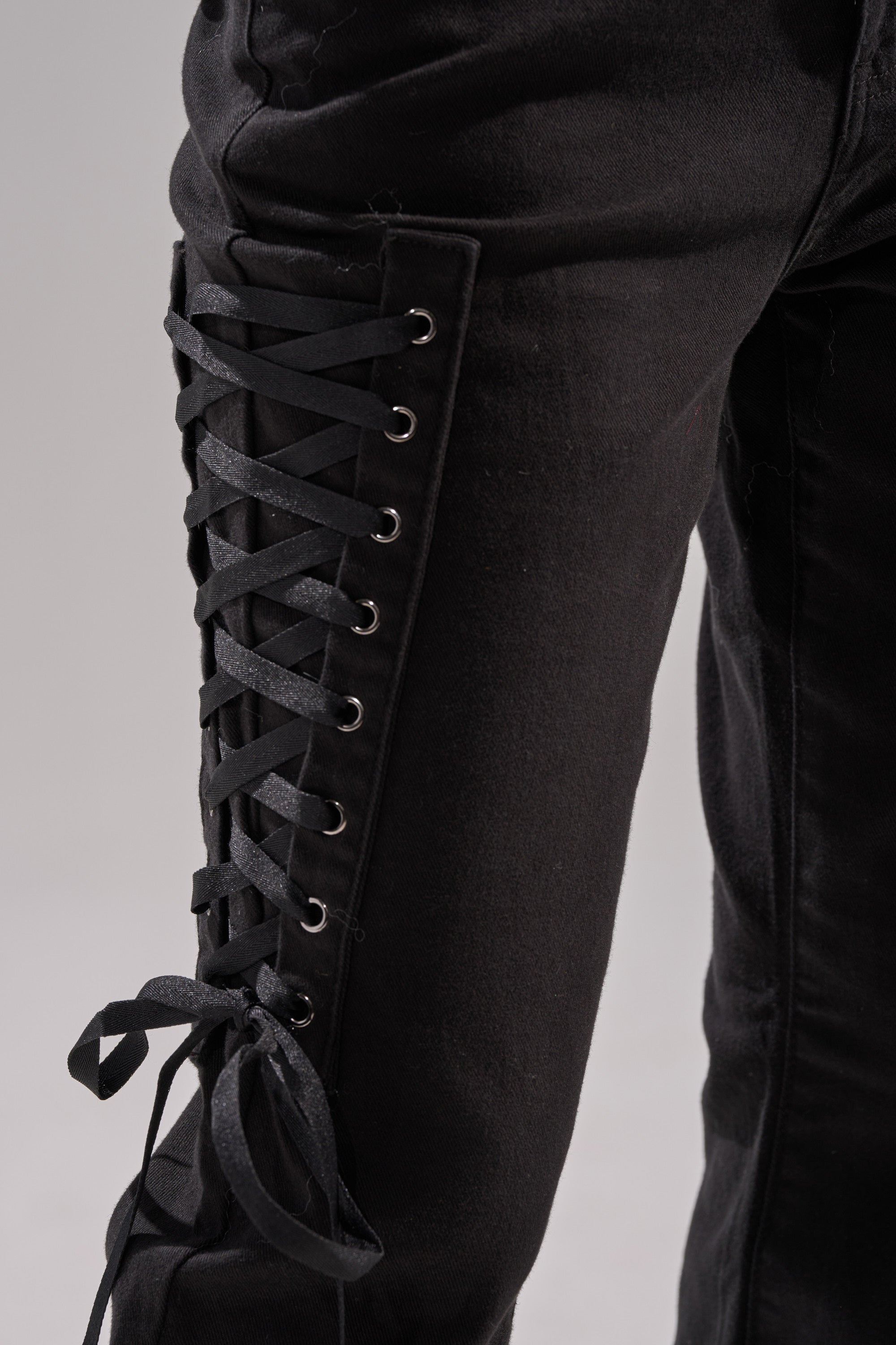 Close-up of LOW LIFE LACE-UP JEANS in a flared silhouette, with black ribbon laced through metal eyelets along the outer thigh and tied at the knee. The background is neutral and softly blurred.