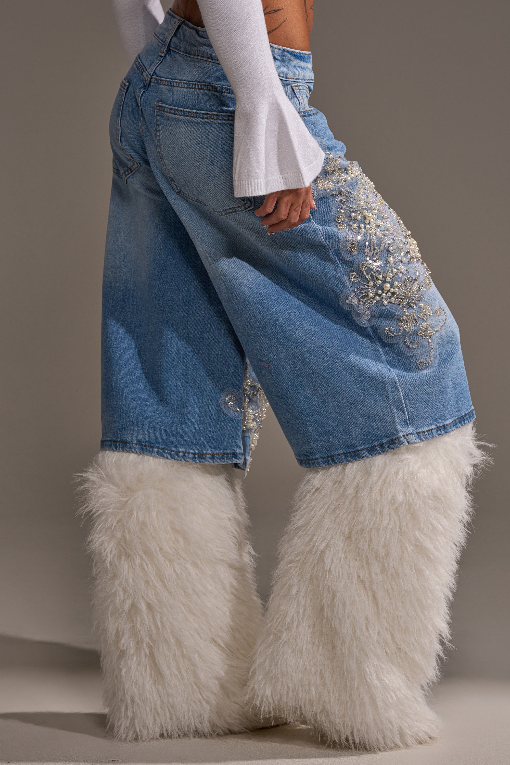 A person wears MADDIE EMBELLISHED BERMUDA SHORTS featuring ornate embroidery and white faux fur accents, paired with a white long-sleeve top, standing against a neutral background.
