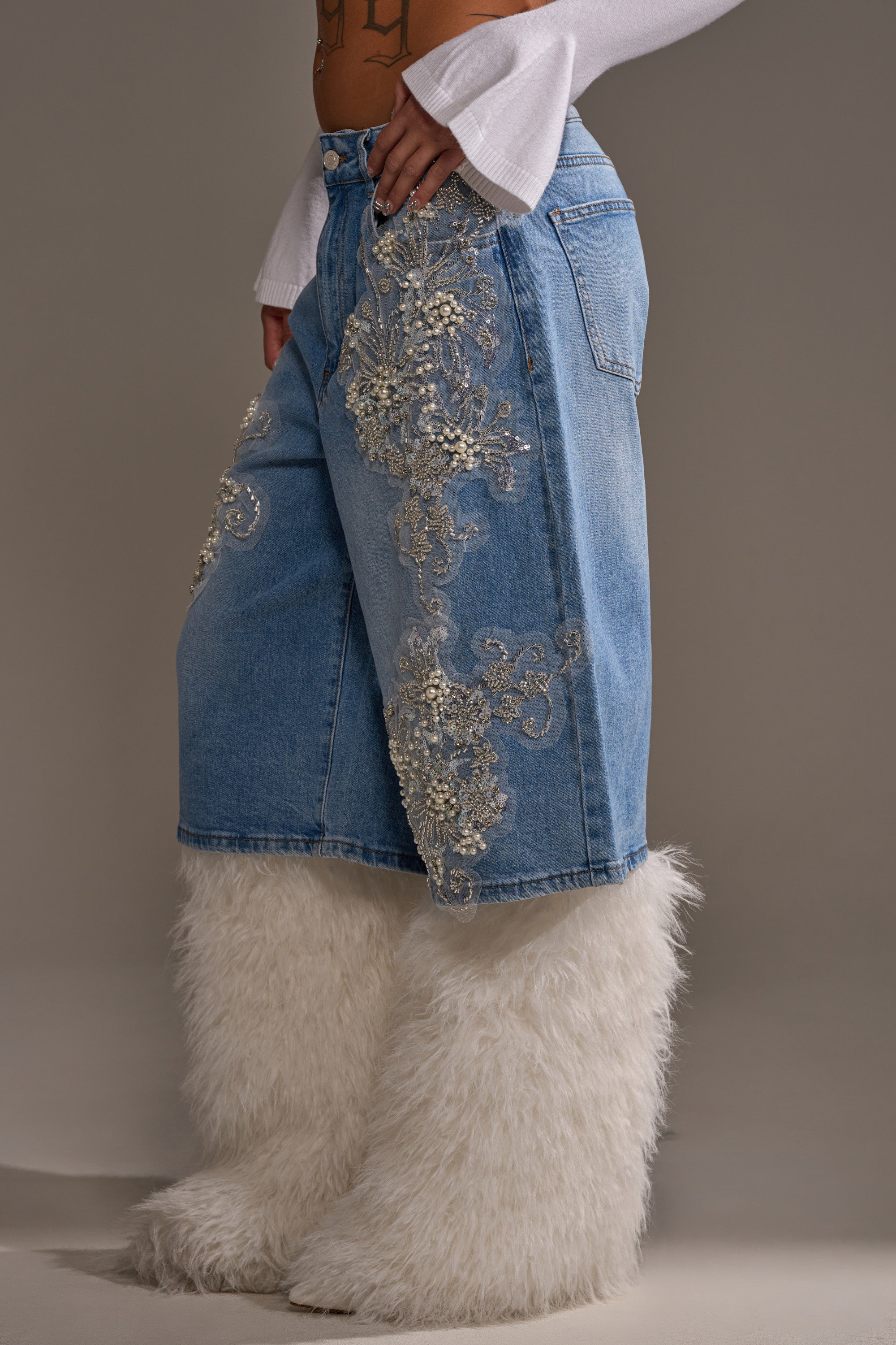 A person showcases the MADDIE EMBELLISHED BERMUDA SHORT, featuring light-wash denim with silver embroidery and sequins, styled with fluffy white fur leg warmers. Only their lower body is visible against a plain background.